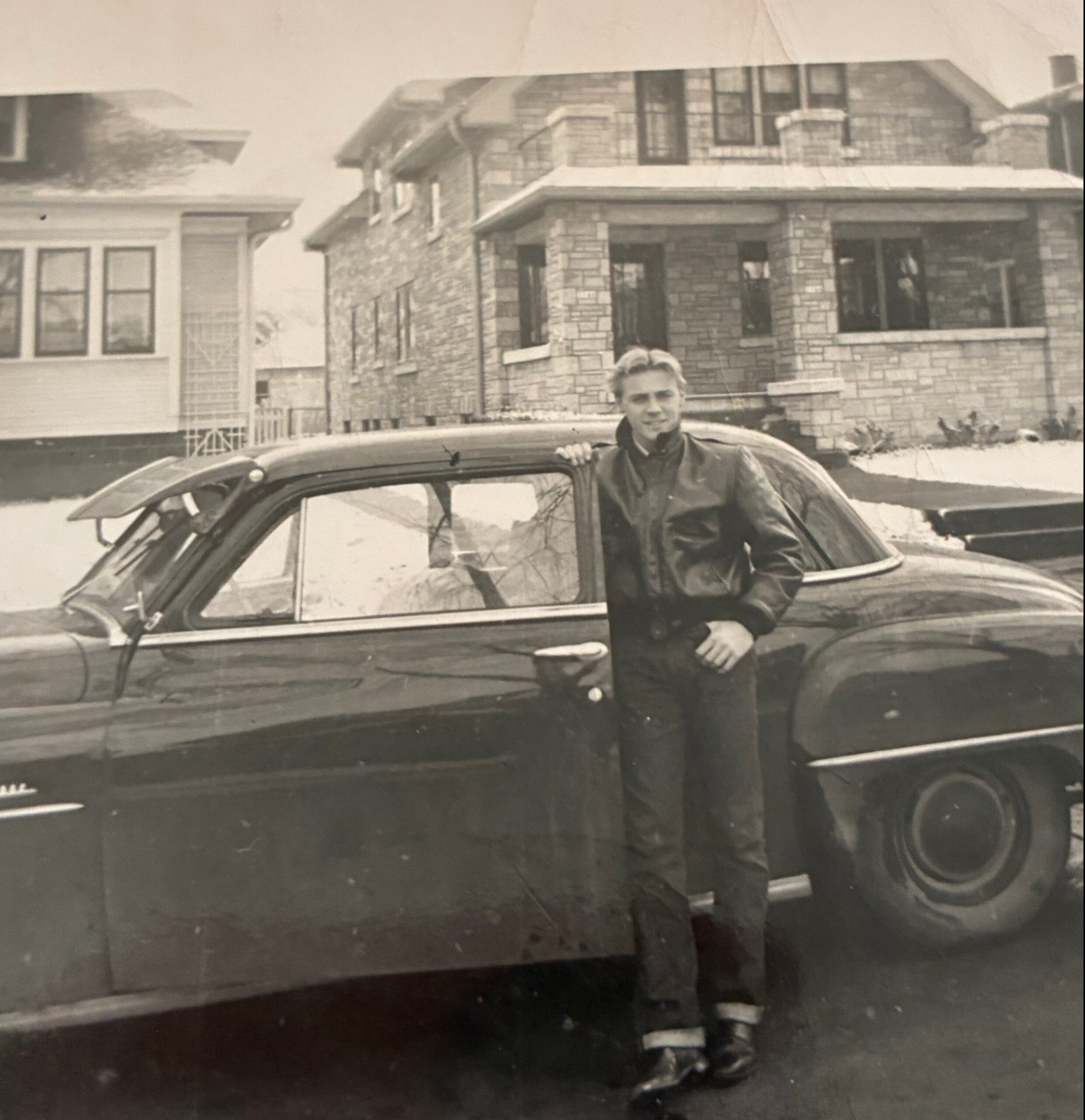 Man leaning on the hood of a vintage car parked on a residential street with stone houses in the background.] [Note: shorten if needed for real usage]