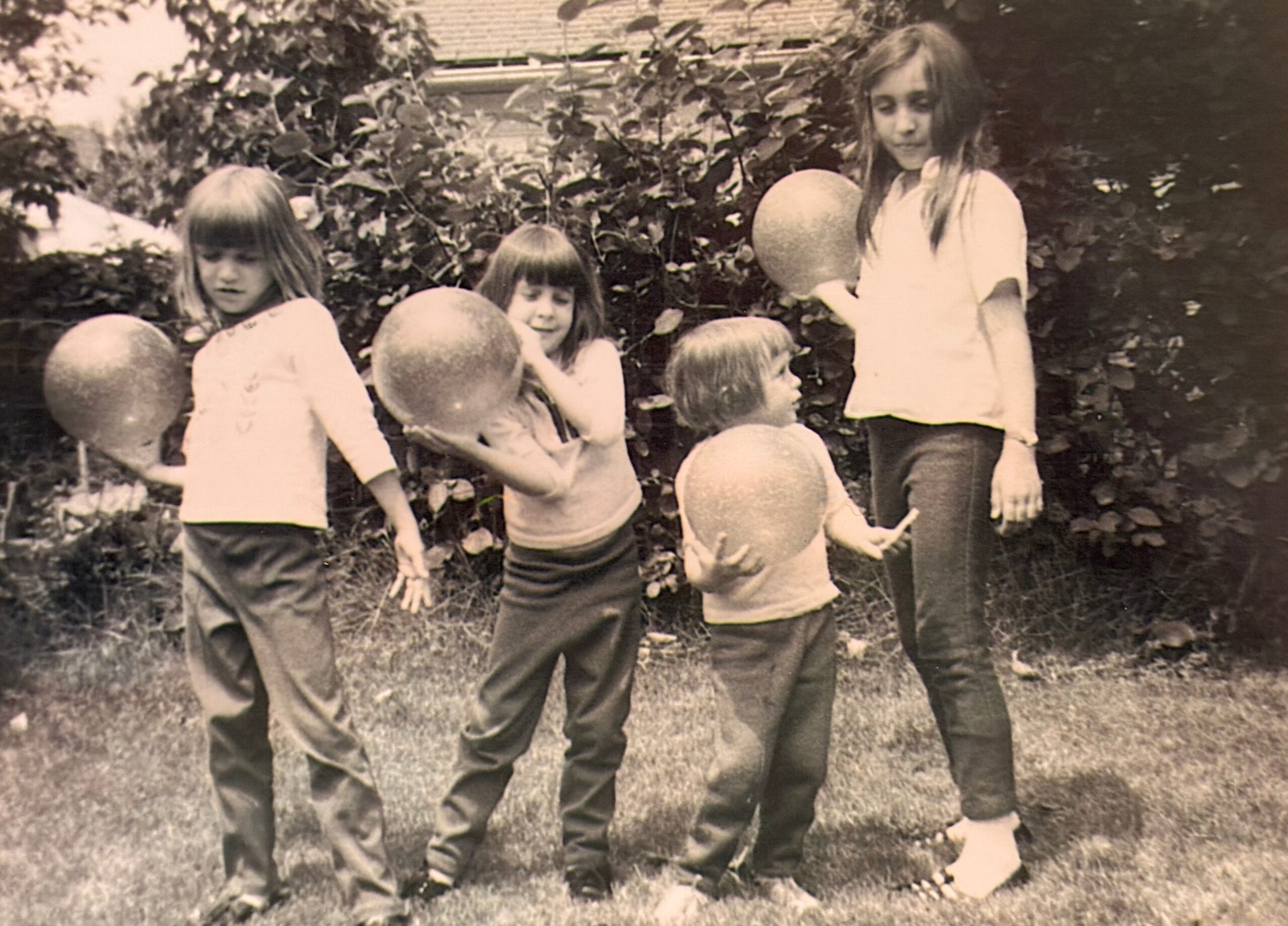 Group of children outdoors, some holding round balloons, standing among bushes in a yard on a sunny day.