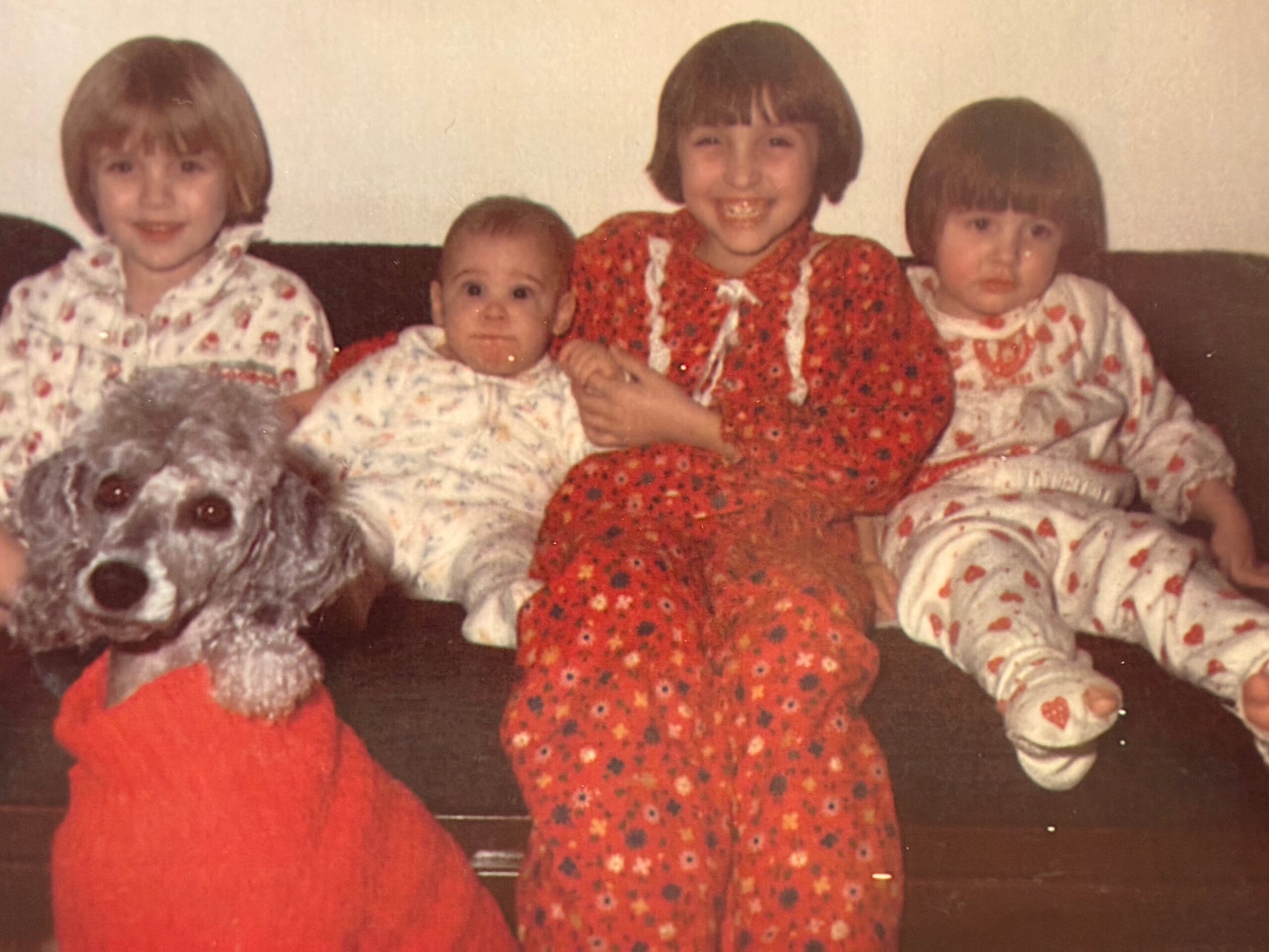 Four young children sit close together on a sofa, wearing matching festive pjs; a large fluffy white dog rests on the left edge of the frame.