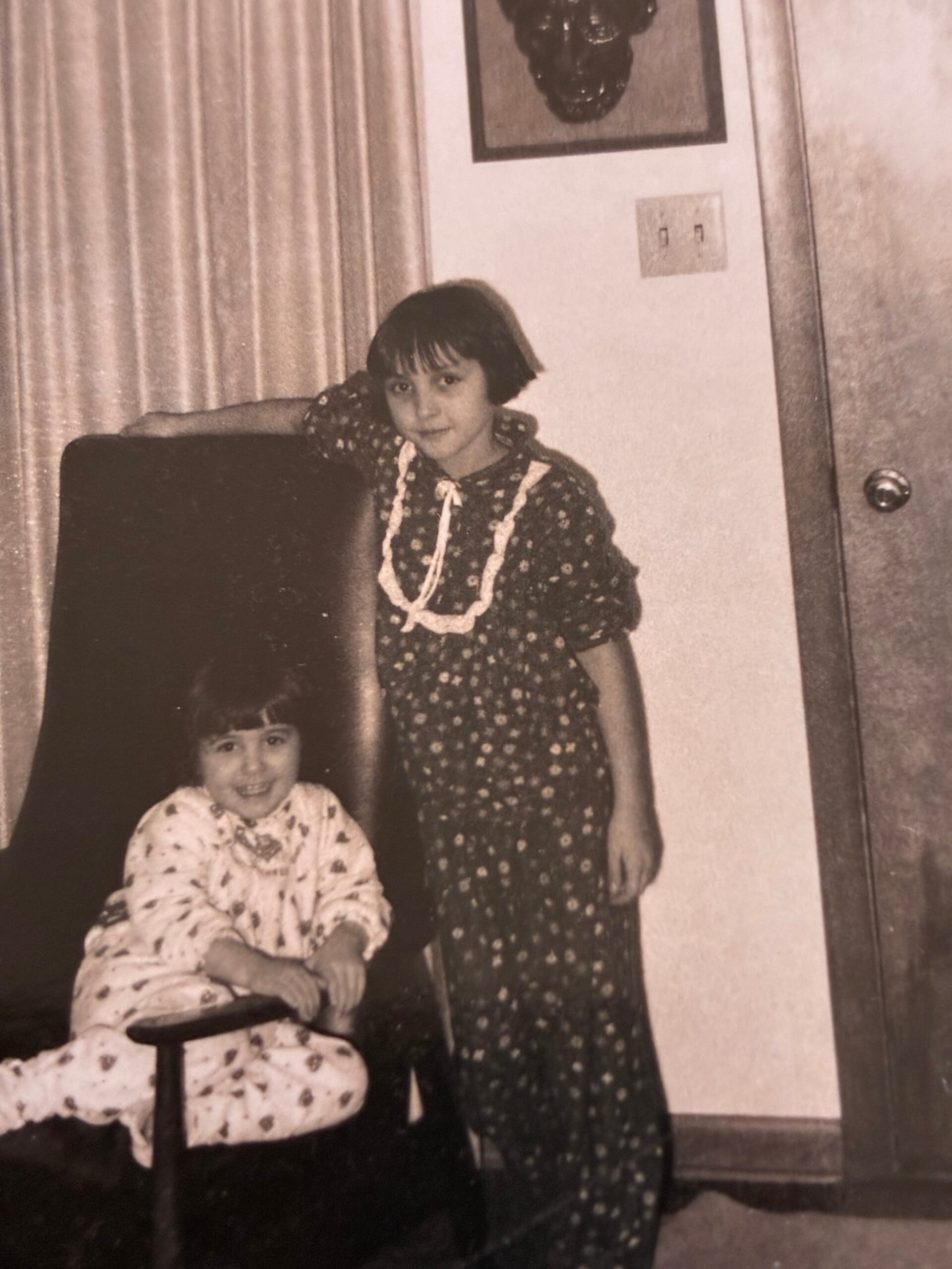 Two young girls pose in a living room: a standing girl in a floral dress with a white necklace beside a seated younger girl in patterned pajamas.