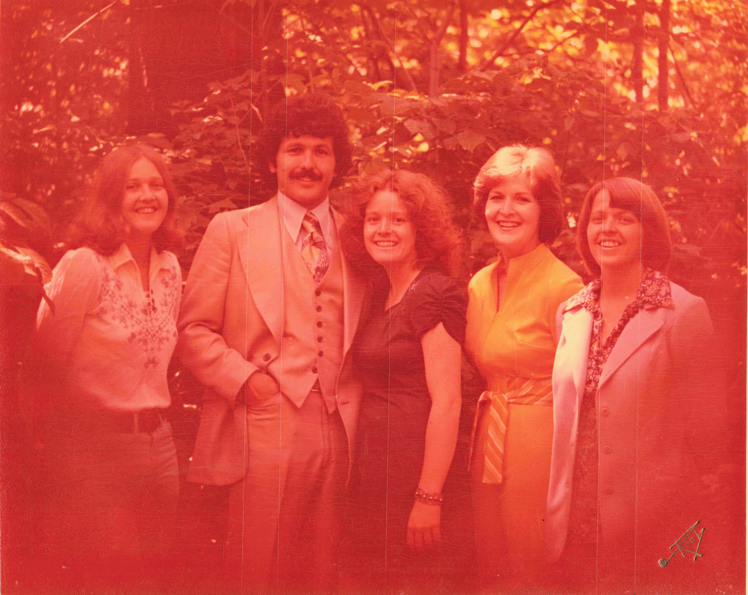 Five adults posing outdoors in a wooded area, smiling, wearing 1970s fashion.875?
