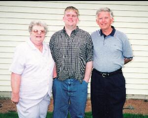 Three people stand outside a white siding house: an older woman in white, a young man in a checkered shirt, and an older man in a blue polo.