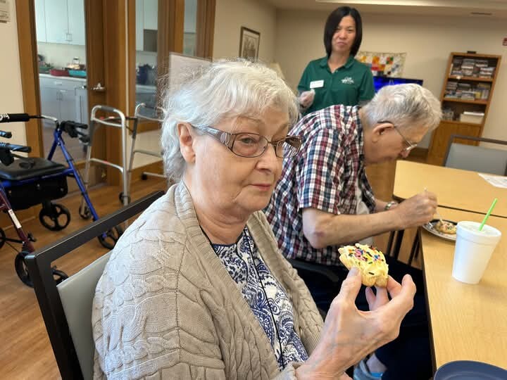 Elderly woman with glasses holding a sprinkled donut at a table in a communal room; a man sits nearby eating and a caregiver stands in the background.