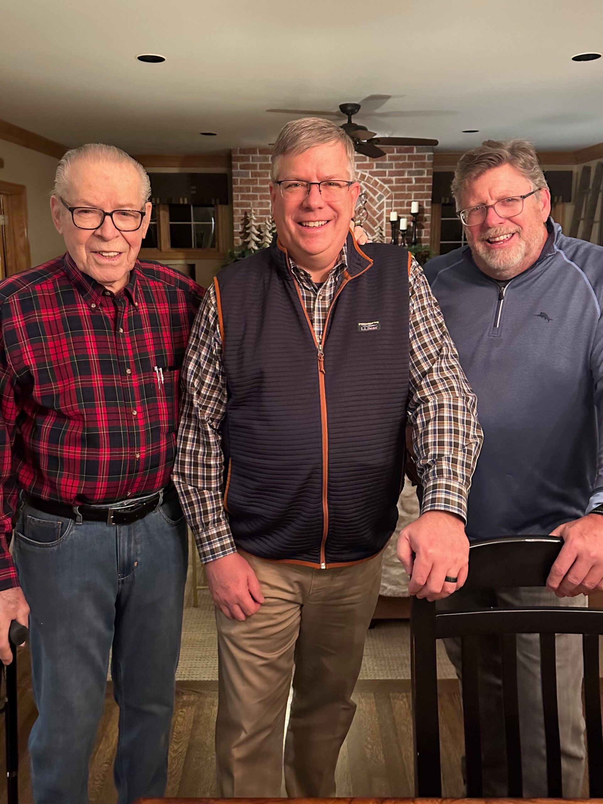 Three men pose for a photo indoors, smiling in front of a brick fireplace; left man in red plaid shirt, center in vest and plaid shirt, right in blue zip top.