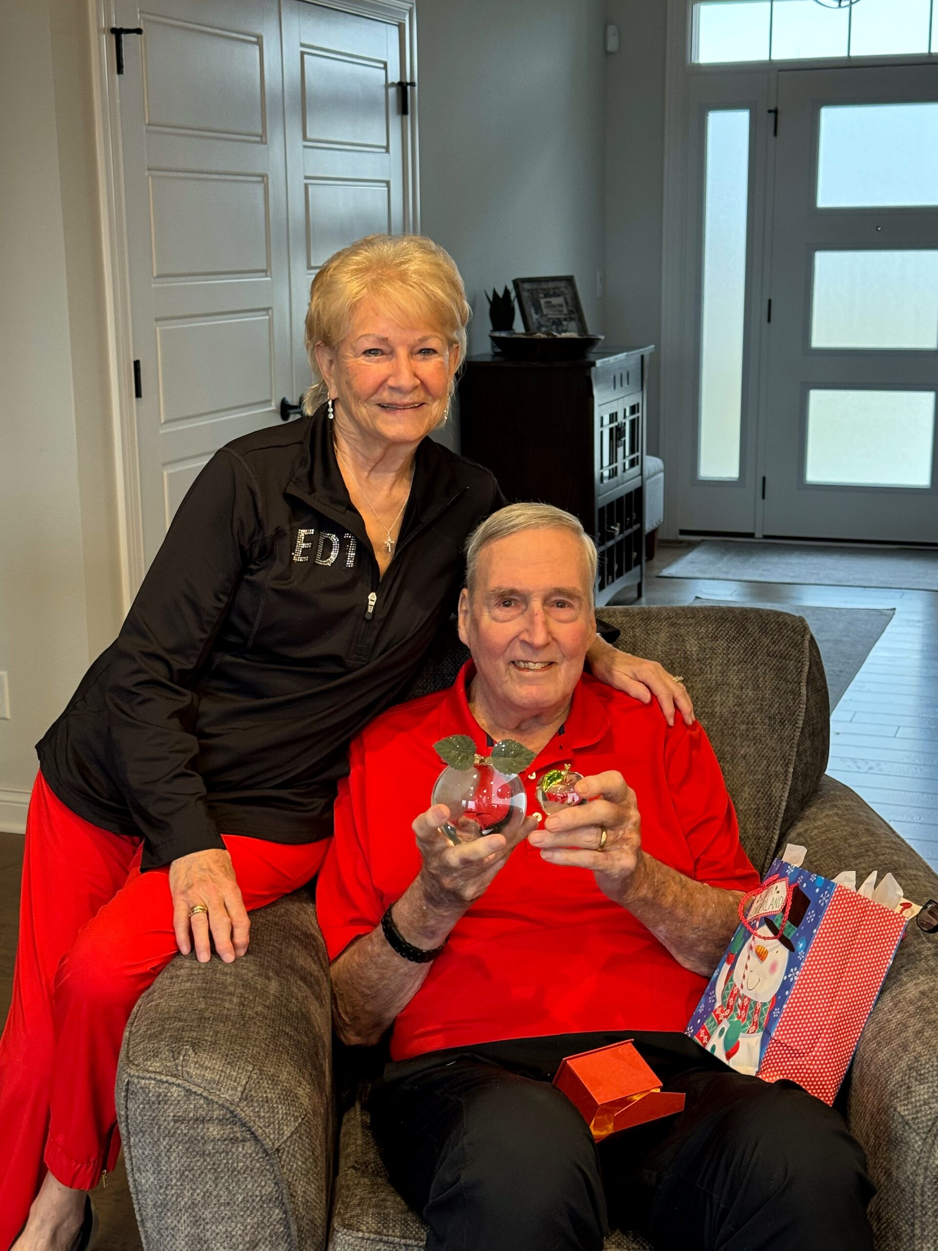 Two elderly women smiling in a living room: one stands beside a seated friend, both dressed in red, holding two clear glass ornaments for a holiday gift.