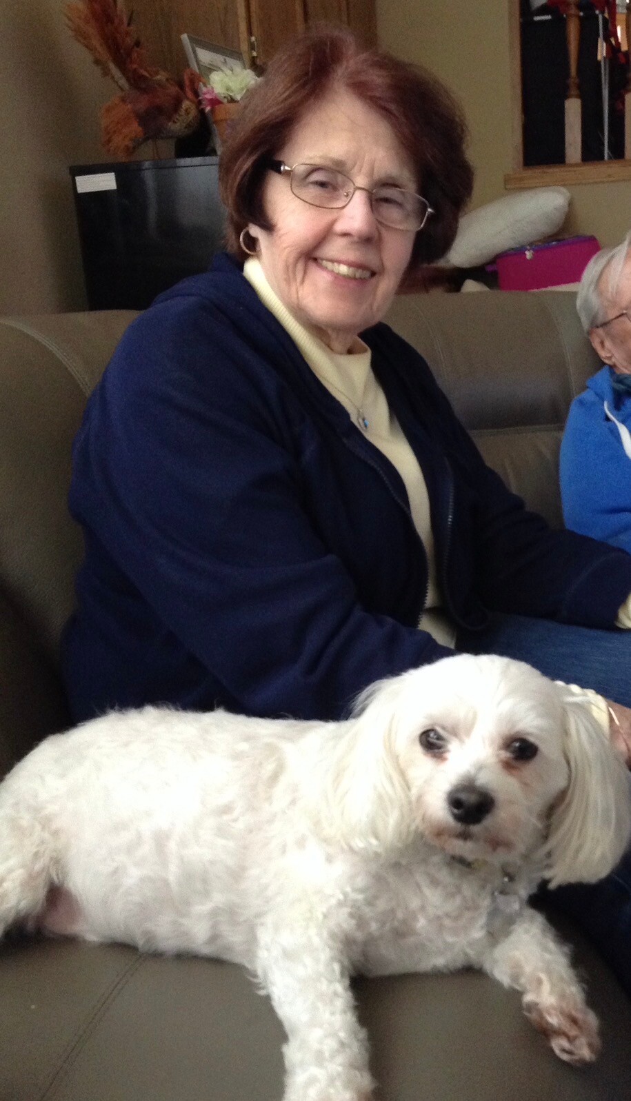 Smiling older woman in glasses sits on a couch with a small white dog lying in front of her on the sofa at home.