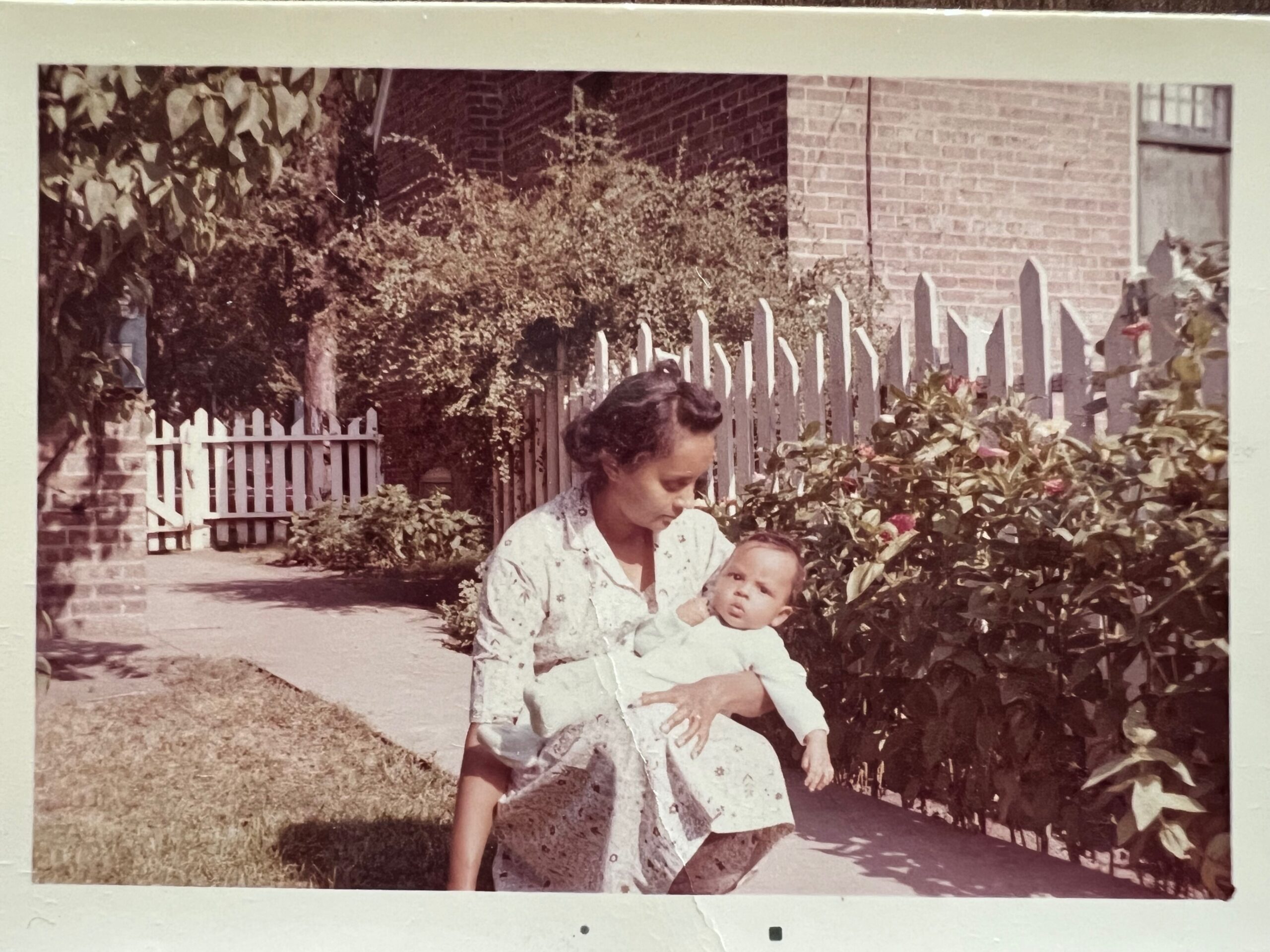 A woman holds a baby in a garden next to a white picket fence and brick building, with bushes and leaves surrounding them.