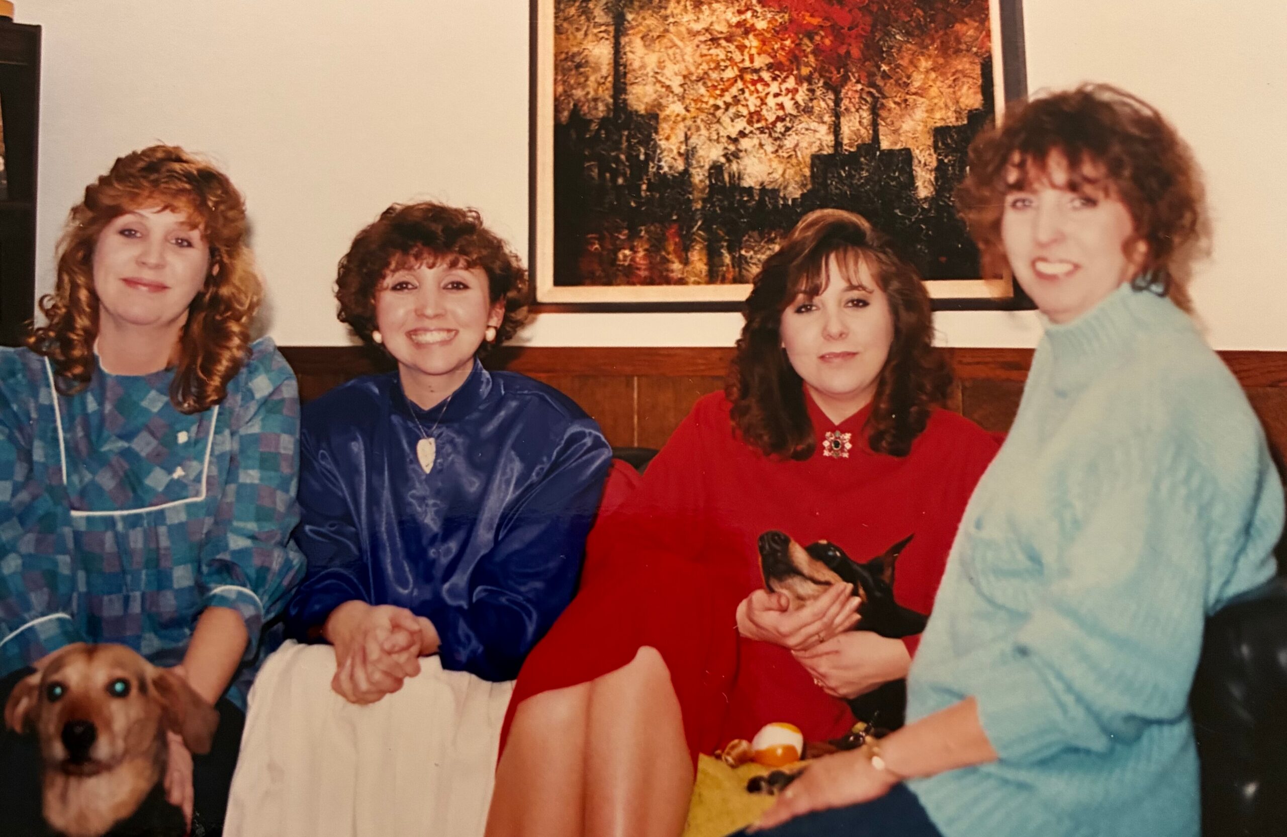 Four women pose together on a couch with a painting on the wall behind them; a brown dog sits at the bottom left of the frame.