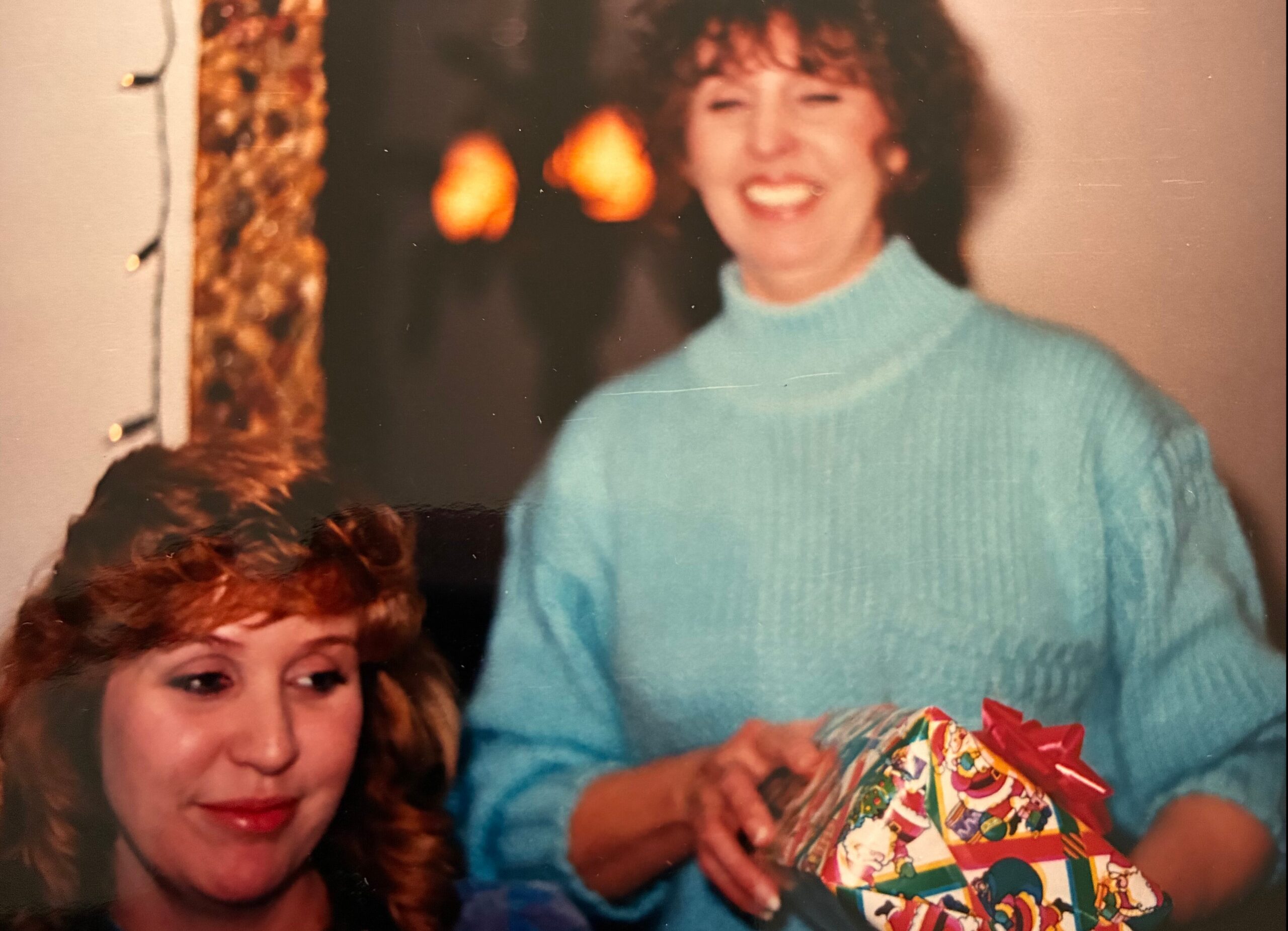 Two women at a holiday gathering; one in a turquoise sweater holds a colorful wrapped gift and smiles, while the other looks on, in a cozy room with string lights nearby.