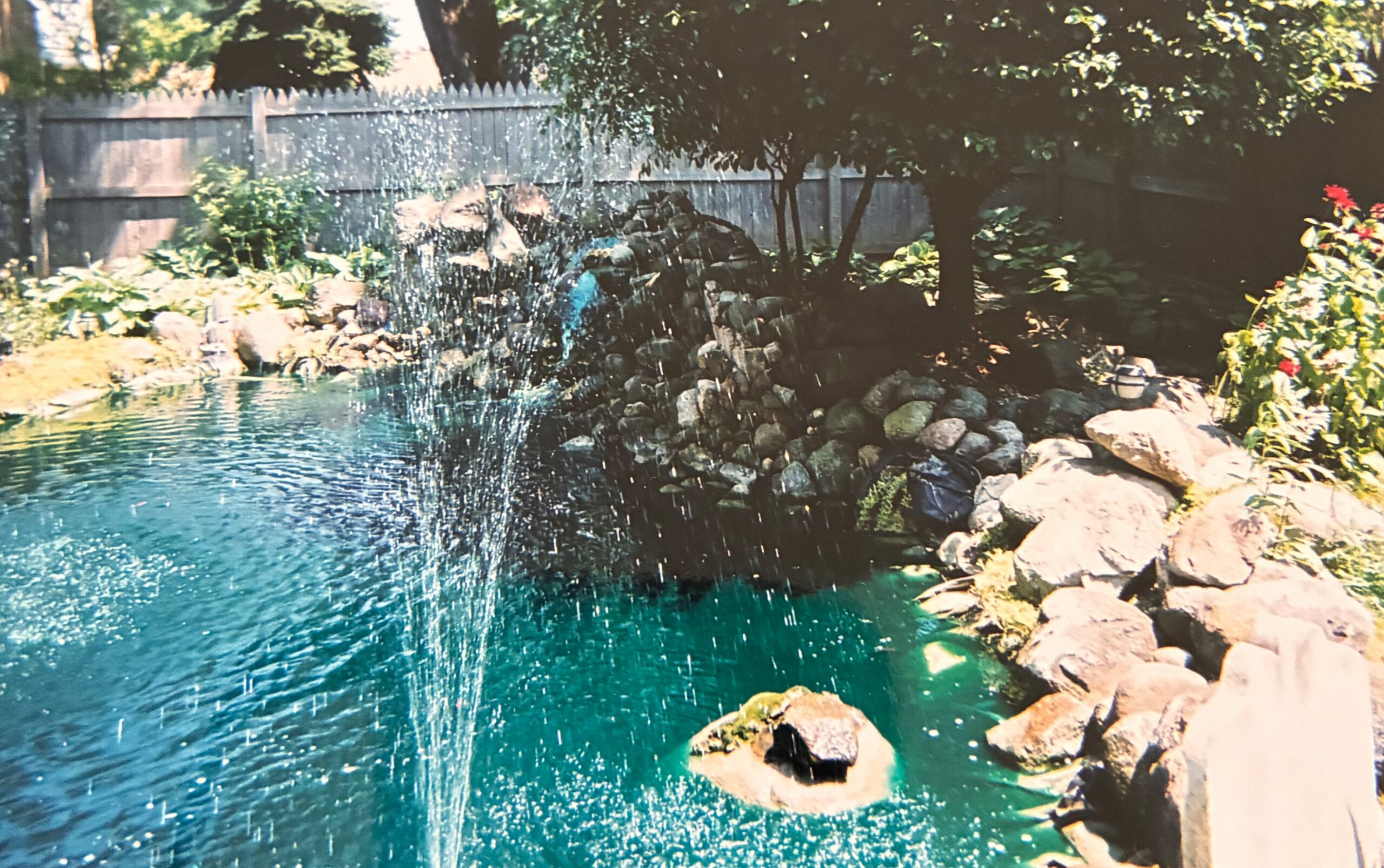 Backyard pond with a rock border and small waterfall cascading into turquoise water, wooden fence in the background and trees casting shade.