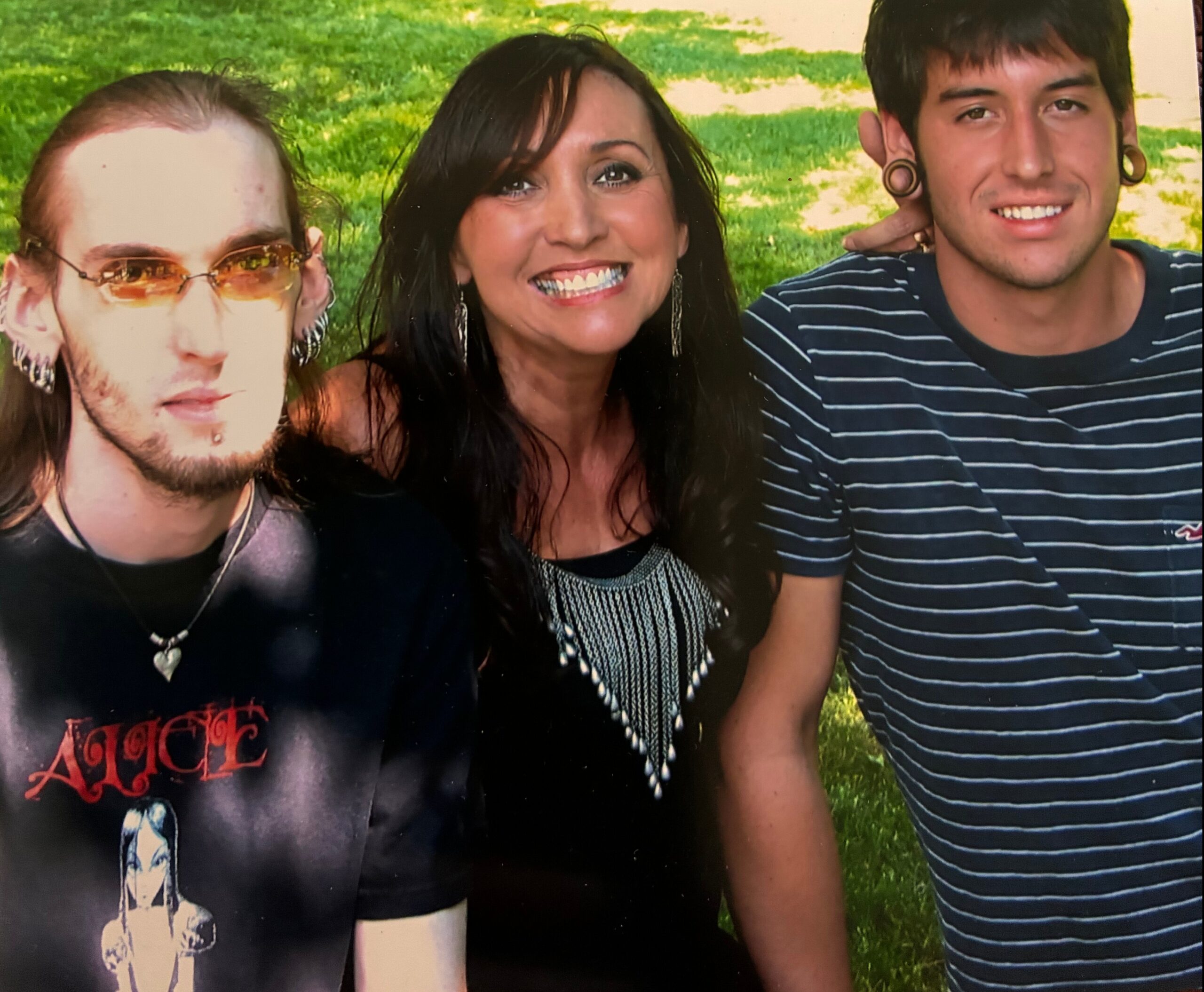 Three friends posing closely for a photo outdoors on a sunny day, smiling at the camera with green grass in the background.