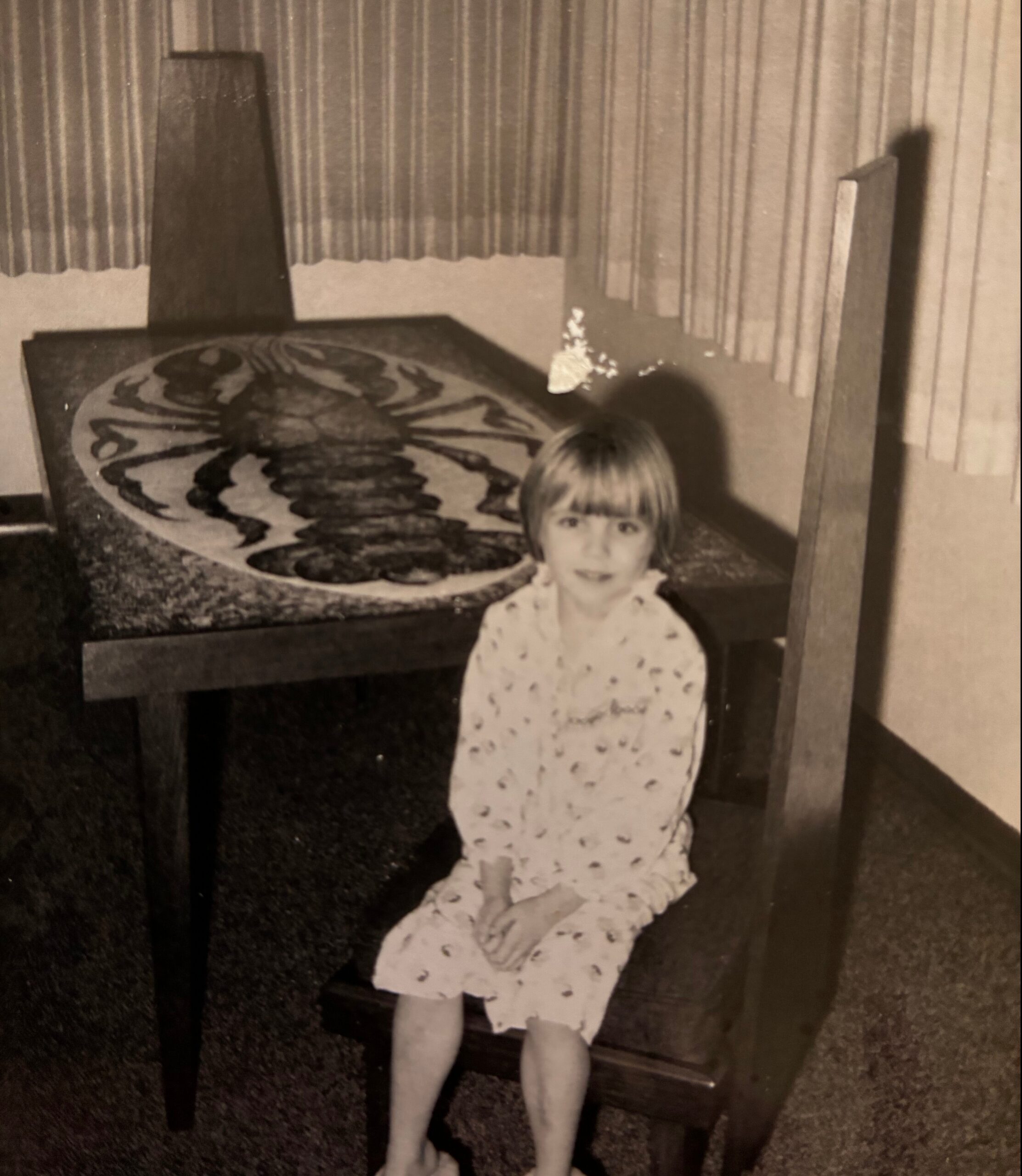 A young girl in a light, polka-dot dress standing in a vintage living room with heavy vertical blinds and a round patterned rug.