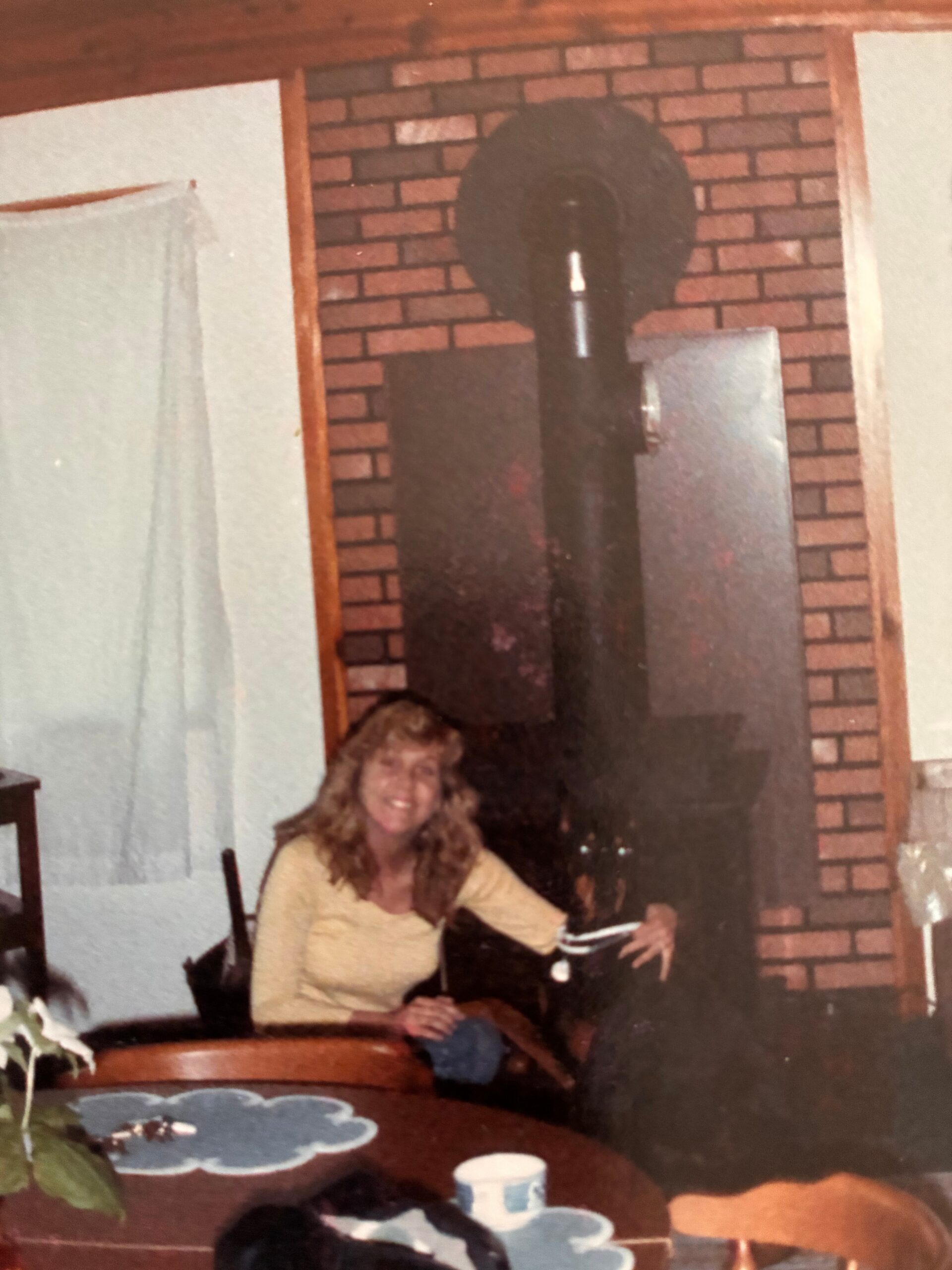 Smiling woman sits at a dining table during a party, with a brick wall and fireplace in the background.