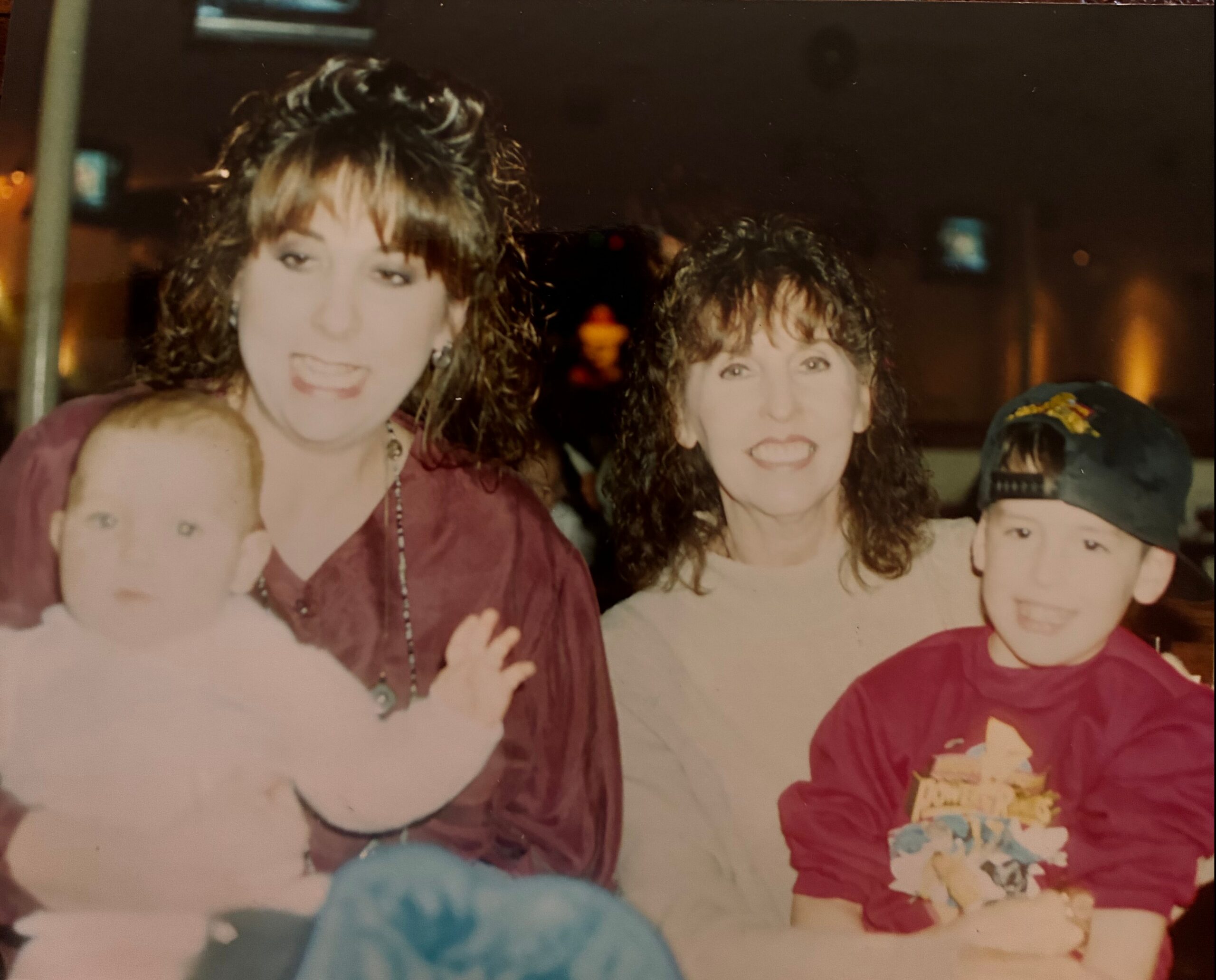 Group of people smiling for a casual photo at night; two women in maroon tops and a child in a cap on the right.