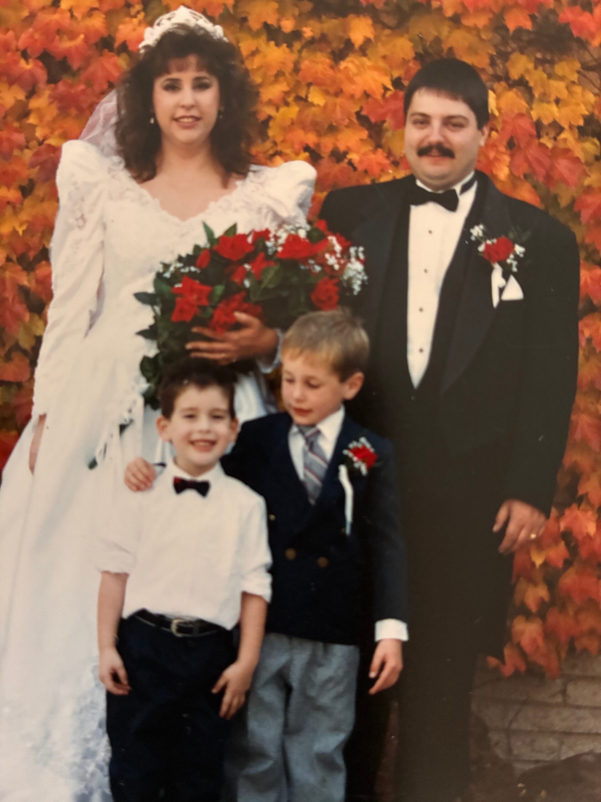 Bride in a white wedding dress holding a red bouquet, standing with a groom in a black tuxedo against a backdrop of orange autumn leaves, plus two children in front.