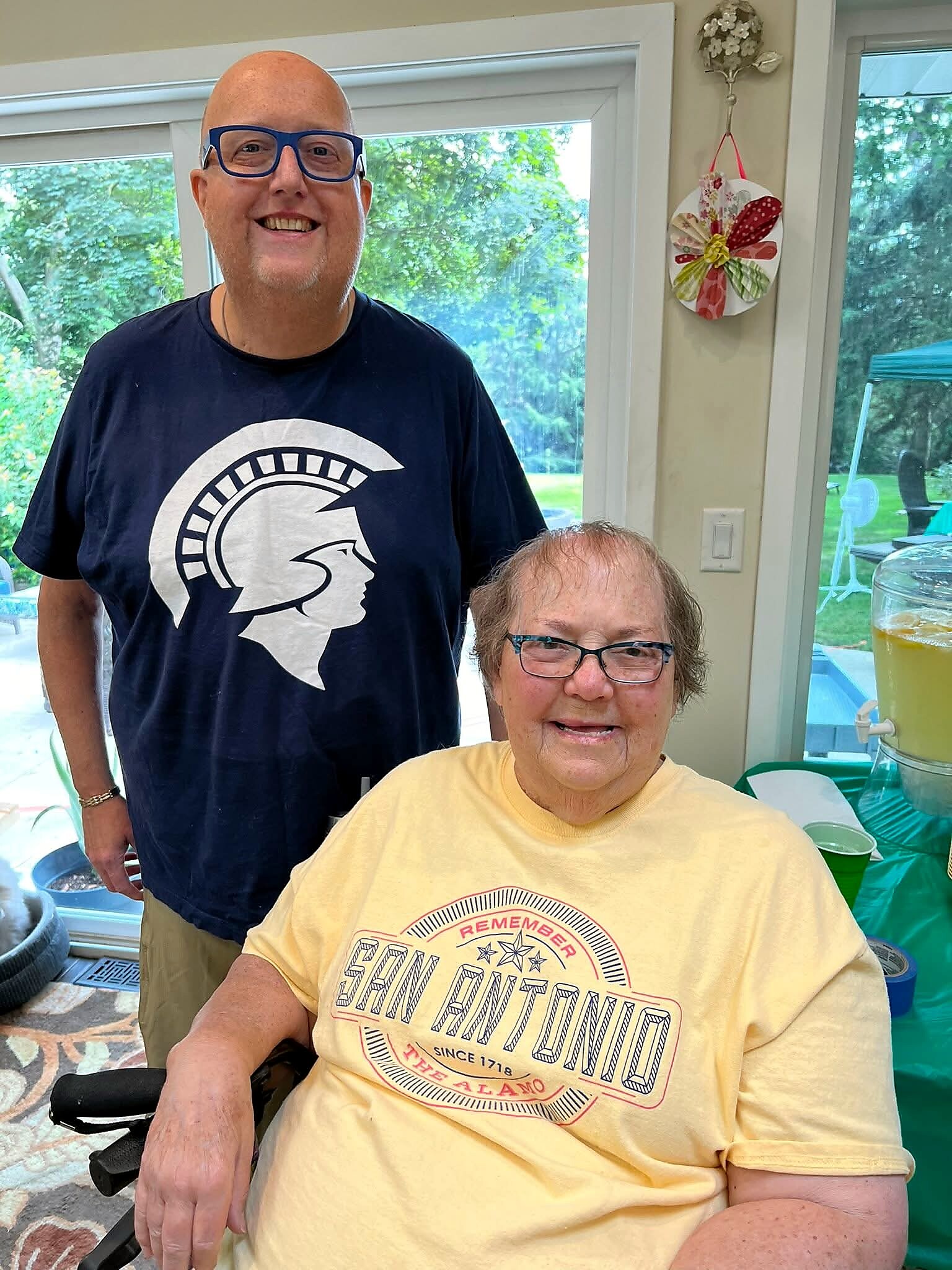 Smiling man in blue glasses wearing a navy T-shirt with a Spartan helmet logo stands beside an elderly woman in a yellow shirt indoors.