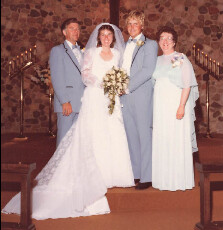 Bride and groom pose with two guests inside a church; bride holds bouquet, all wear light blue or pale attire for a wedding photo