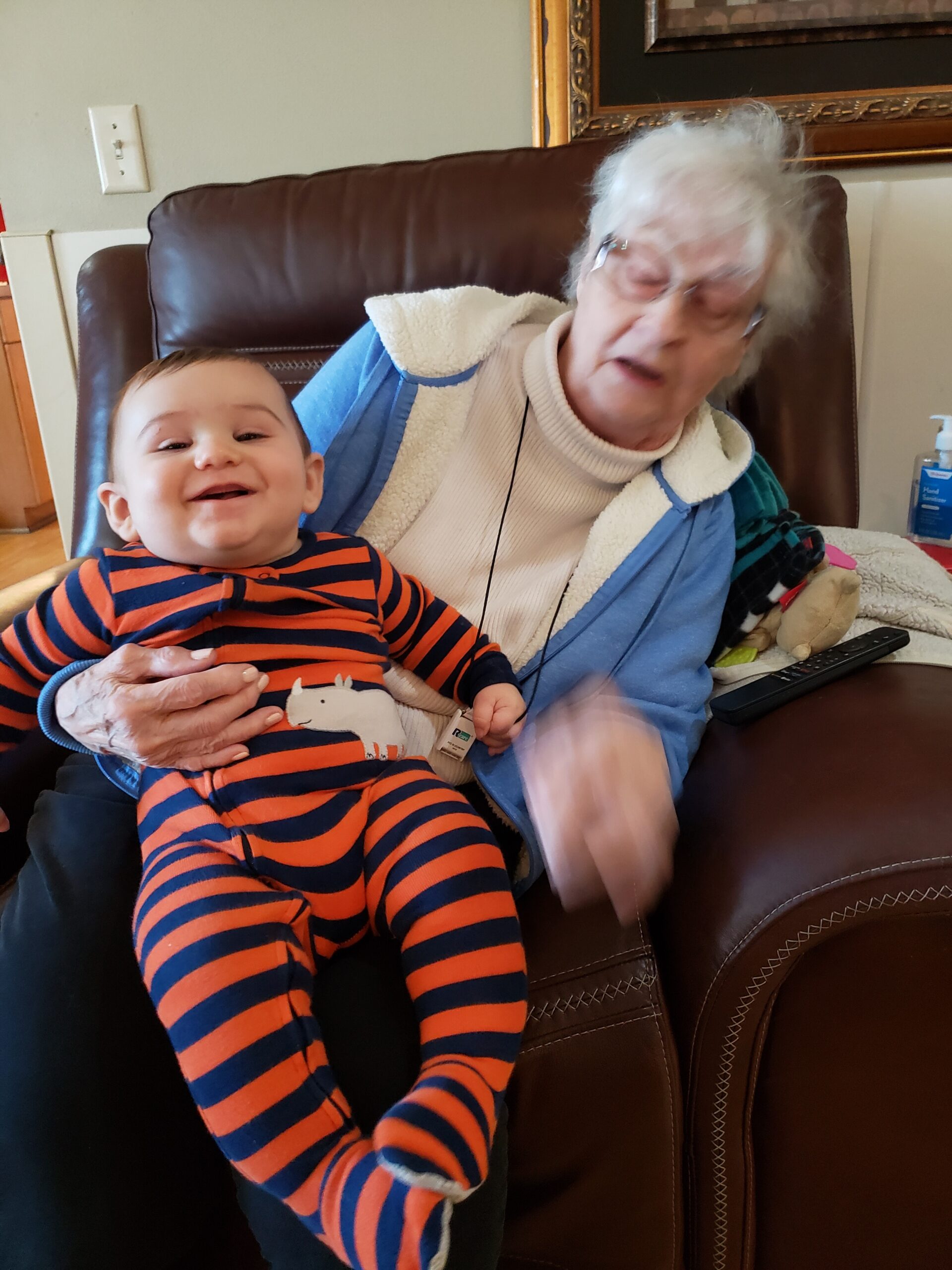 Smiling baby in orange and navy striped onesie sitting on an elderly woman's lap on a brown leather chair indoors.