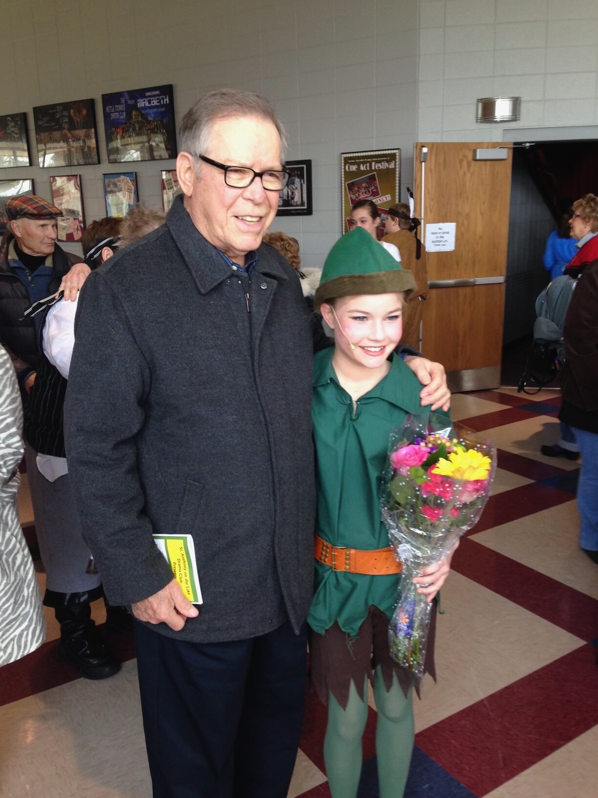Older man in a dark coat posing with a smiling girl in a green elf costume holding a bouquet of flowers at a school/event venue.