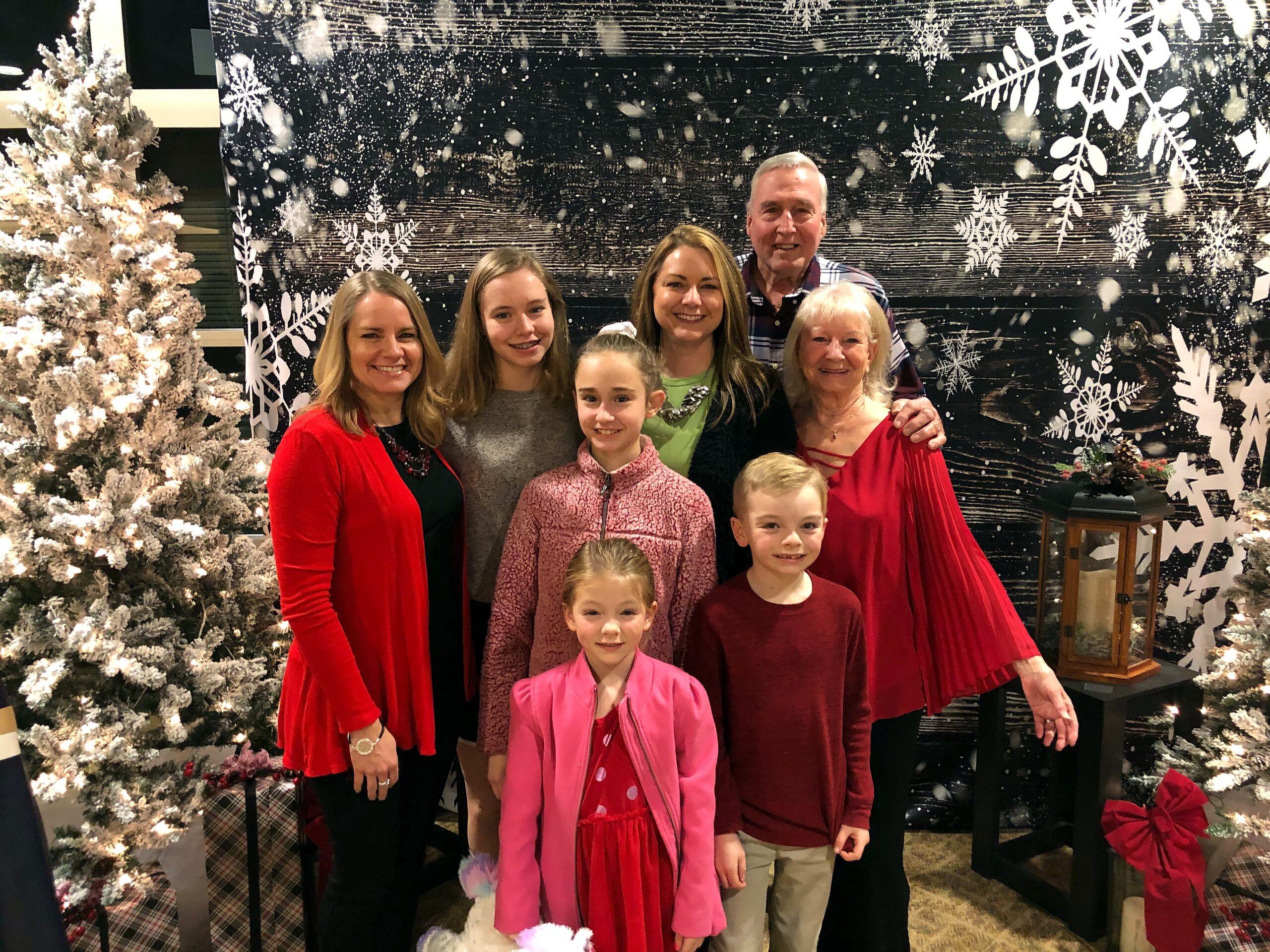 Group of eight people posing together for a holiday photo in front of a snowy black backdrop with snowflakes and frosted Christmas trees on the sides.
