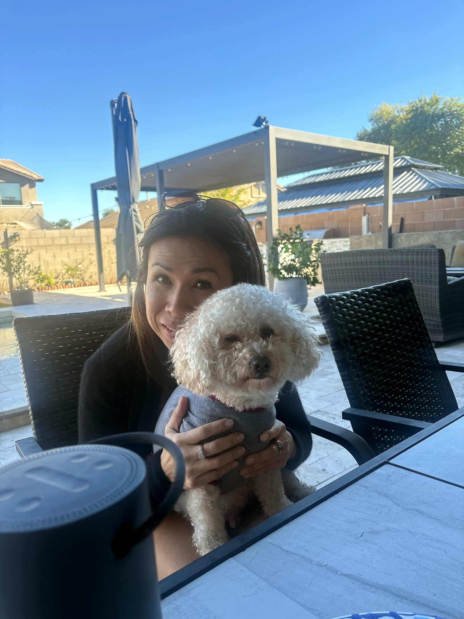 Woman sitting at an outdoor table, holding a fluffy white dog against a bright blue sky.