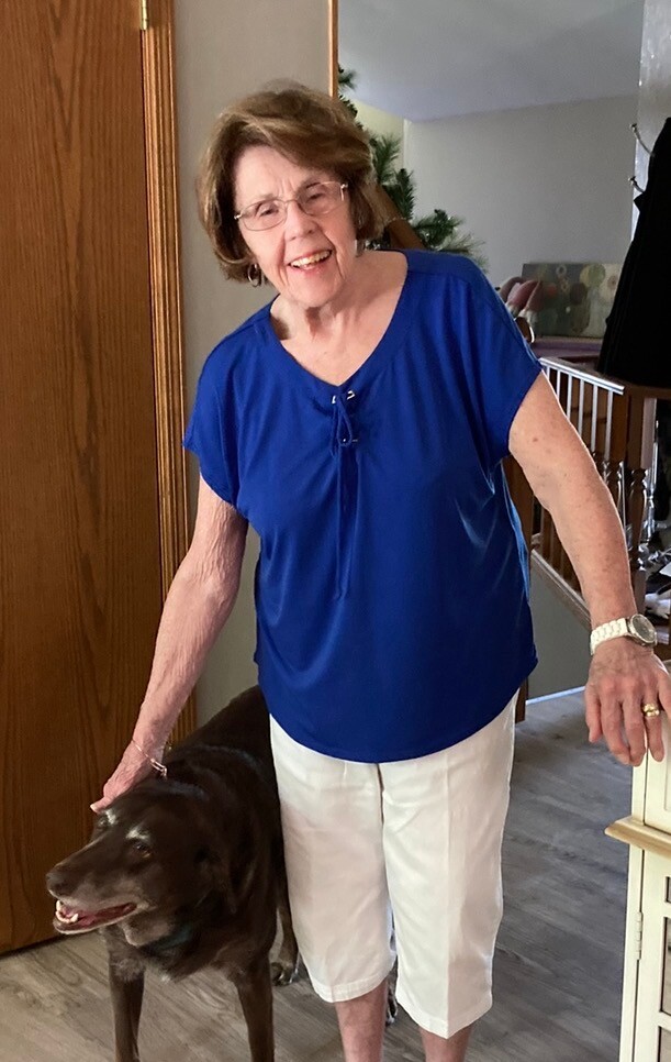 Older woman in a bright blue top and white capri pants smiles while patting a brown dog indoors at home.