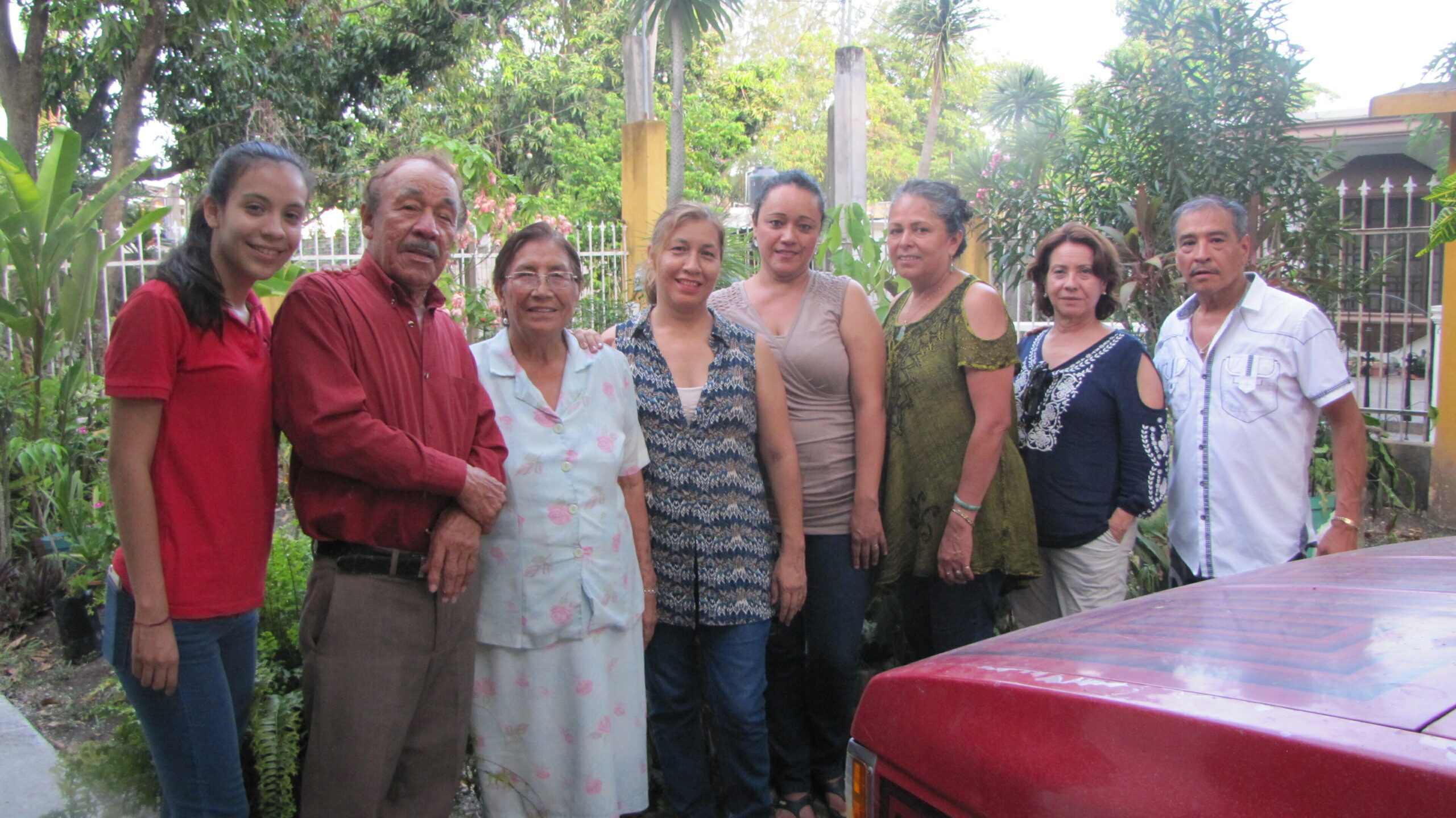 Eight adults pose for a group photo in a lush garden, smiling at the camera by a red car in the foreground.