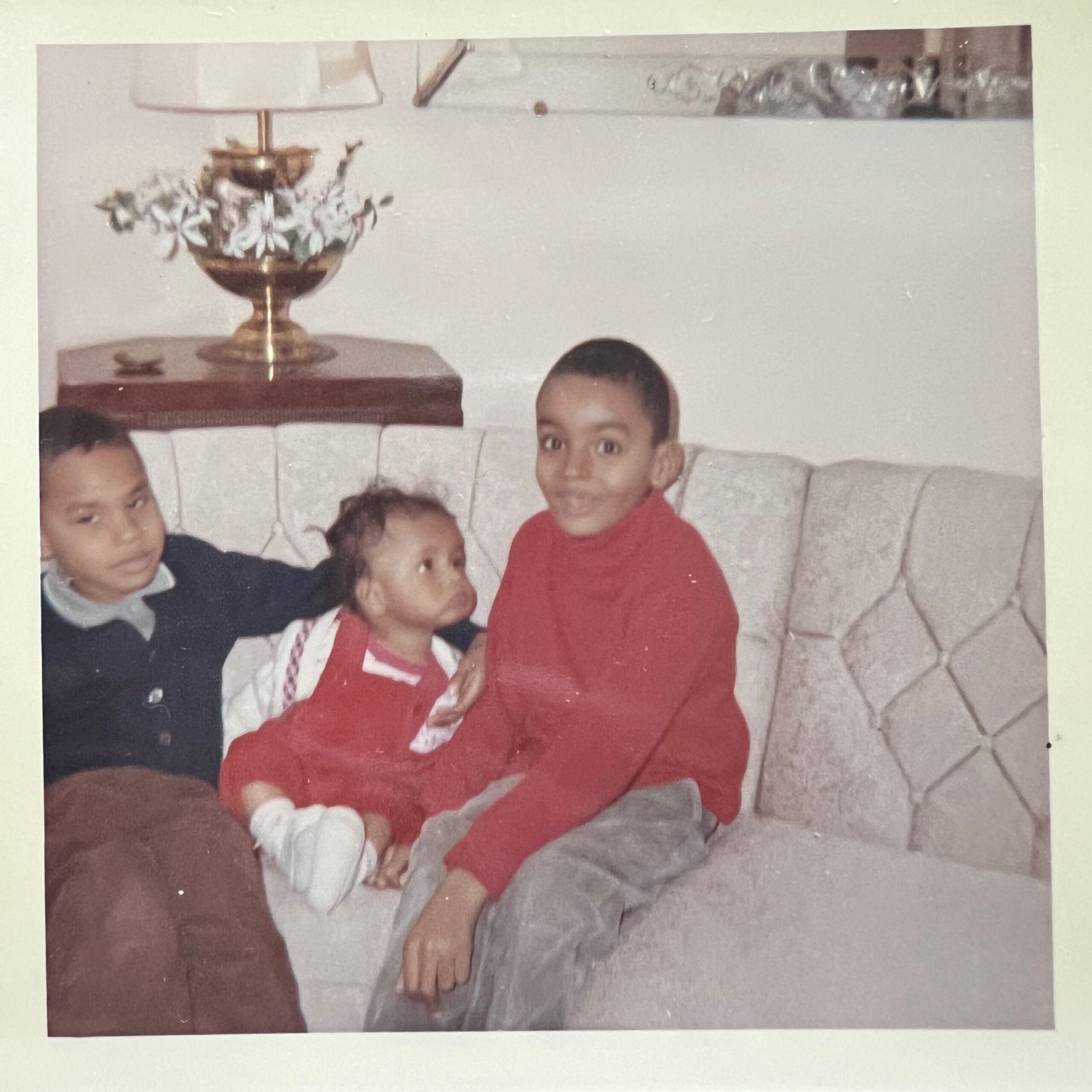 A family group sits on a light-colored couch; children in red outfits, one wearing white gloves, posing for a casual photo with a lamp and decorative vase in the background.