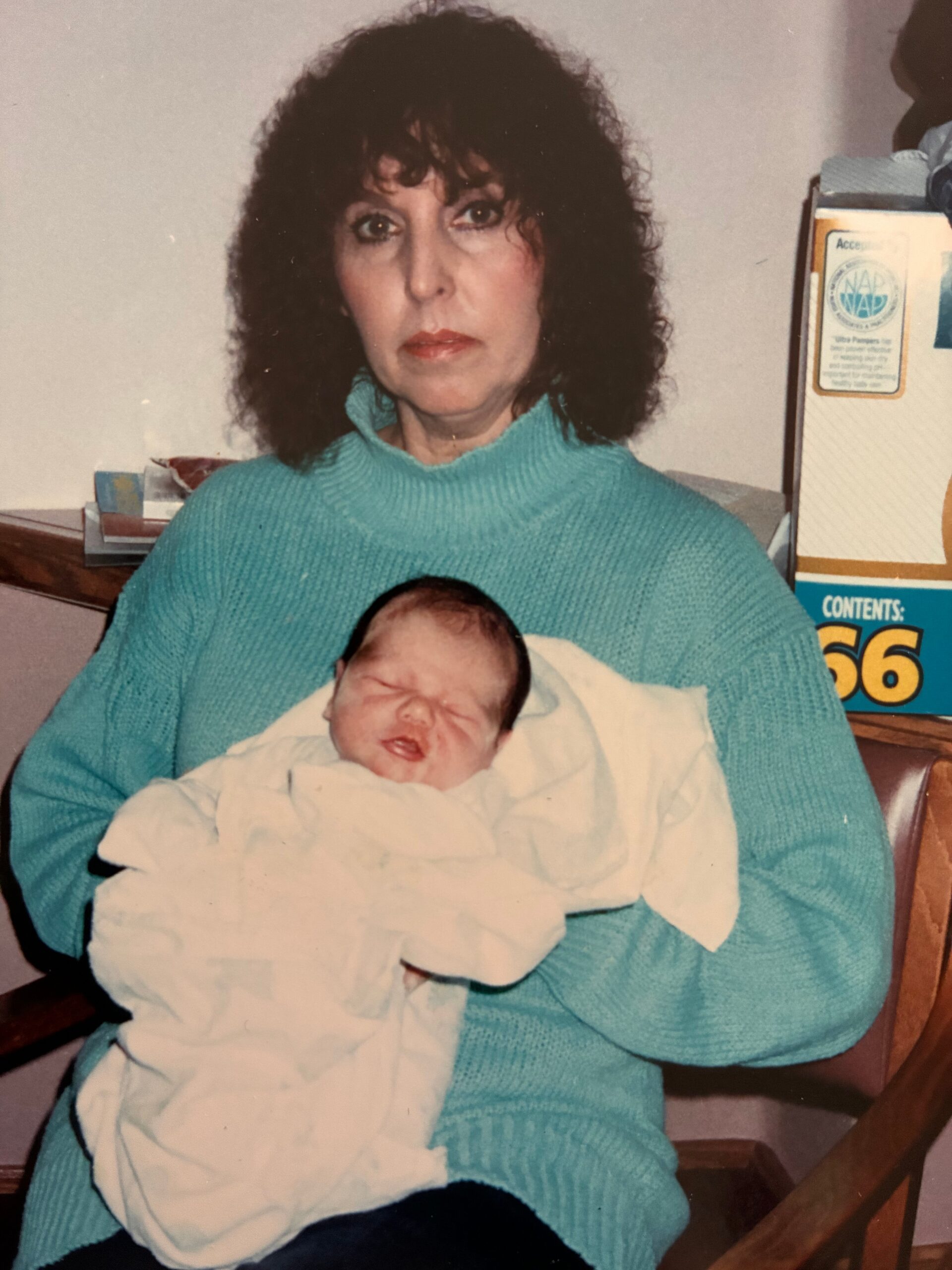 Woman in a teal sweater cradling a swaddled newborn baby wrapped in a white blanket.