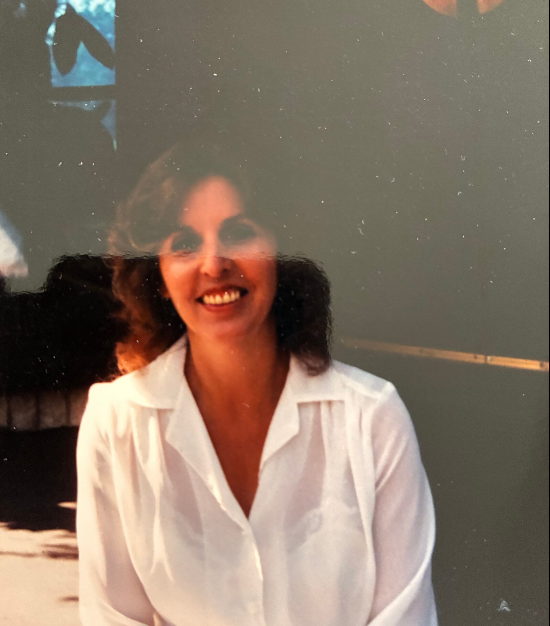 Smiling woman in a white button-up shirt outdoors, dark hair around shoulders, sunlit scene.