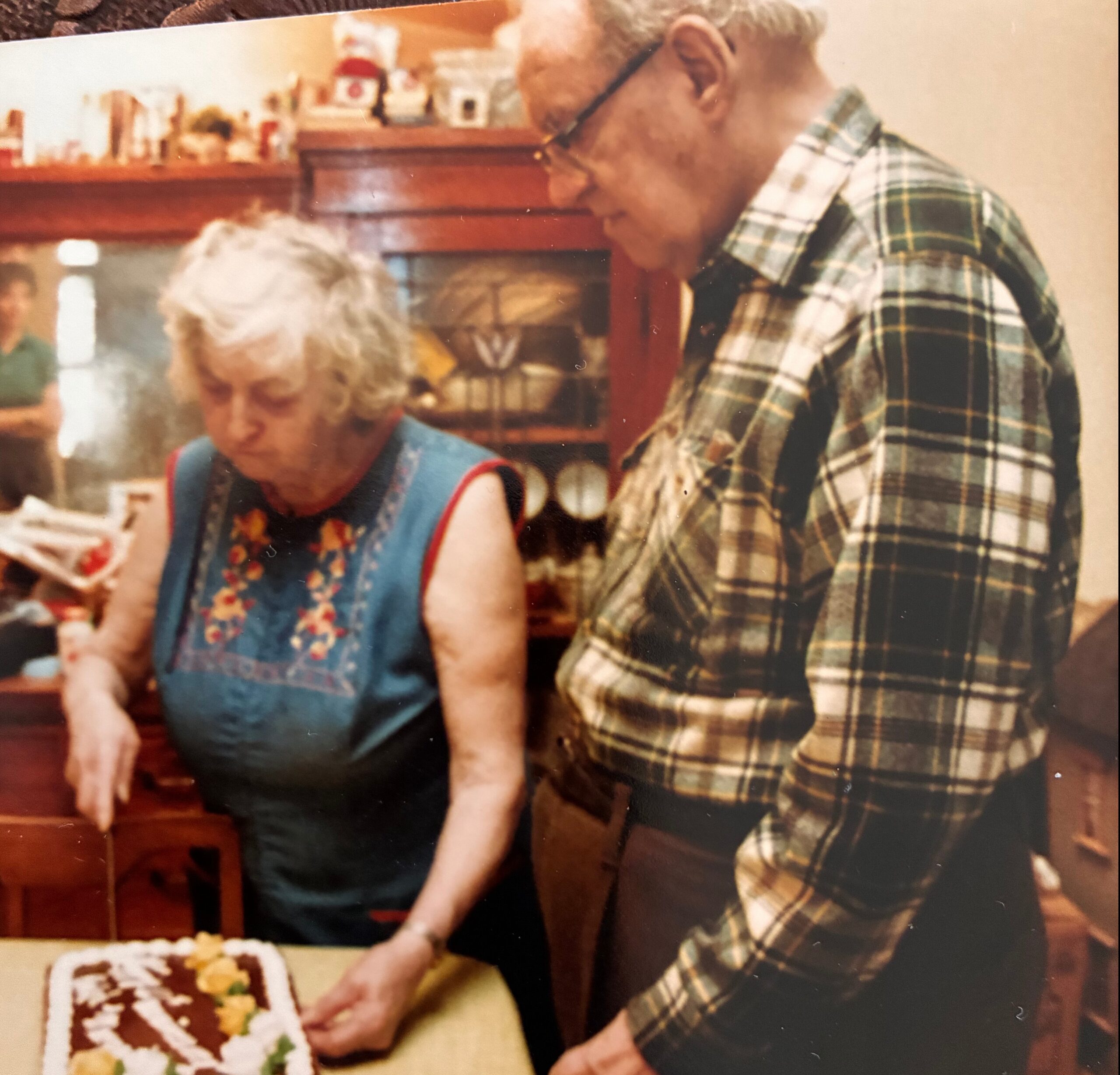 Two elderly people stand by a table, cutting a decorated birthday cake together.