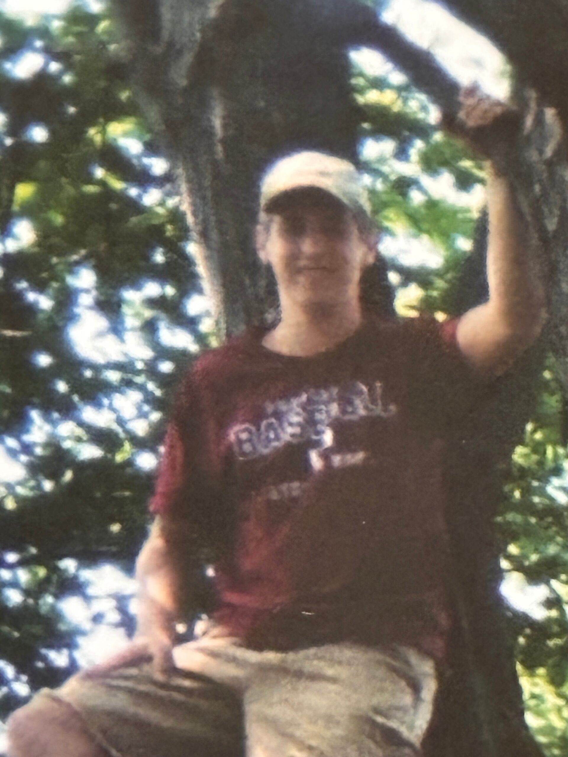 Man wearing a cap sits on a log in a sunlit forest, smiling at the camera.