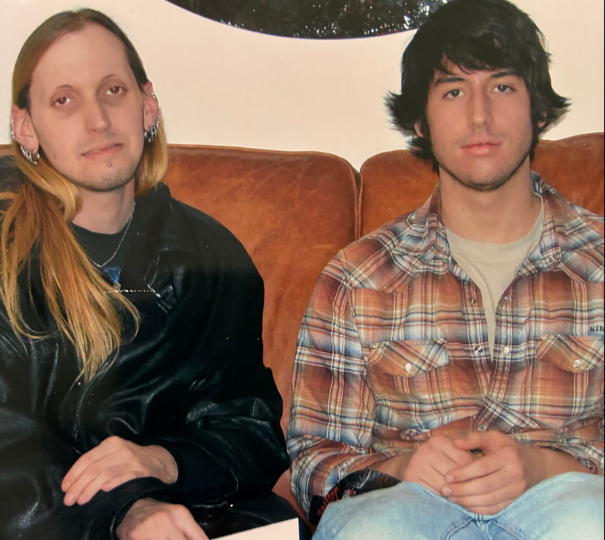 Two people sit on a brown sofa; left person has long blond hair and facial piercings, right person wears a plaid shirt and has dark hair.
