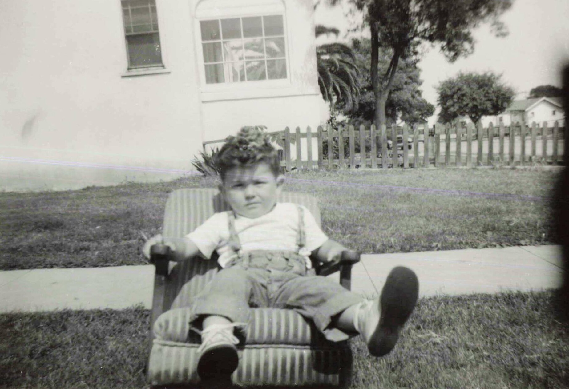 Young child sitting in a striped outdoor chair on a lawn, with a white house, window, and wooden fence in the background