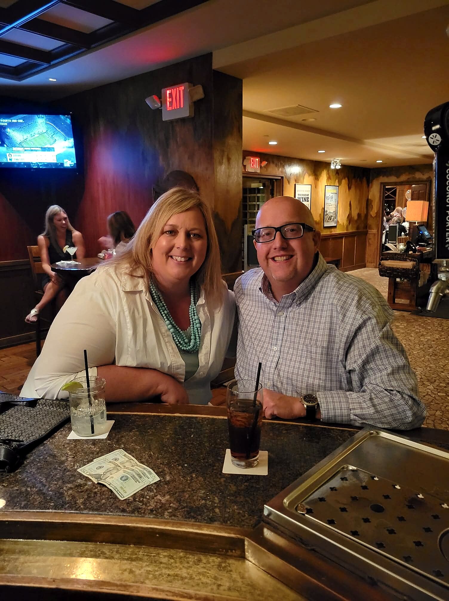 Two people smiling at a bar counter with cocktails in front of them, money on the counter nearby, warm wood bar interior in the background.