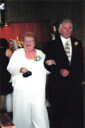 Elderly couple walking arm-in-arm at a formal event; woman in a white pantsuit with a yellow-blue corsage, man in a dark suit.