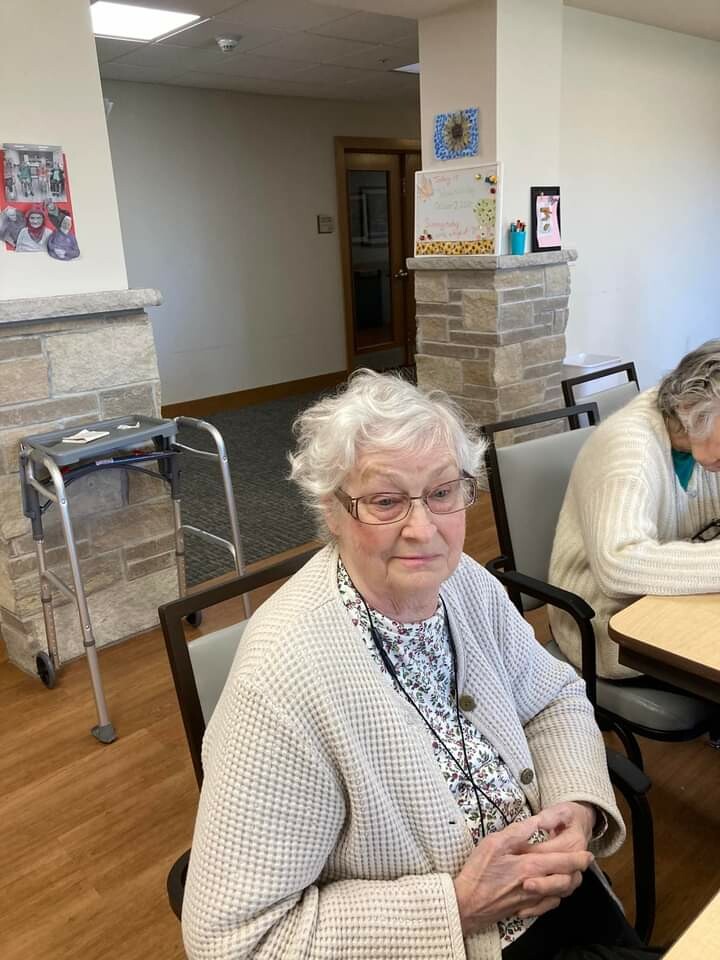 Elderly woman with white hair and glasses sits at a table in a communal room; a walker rests against a stone pillar nearby.