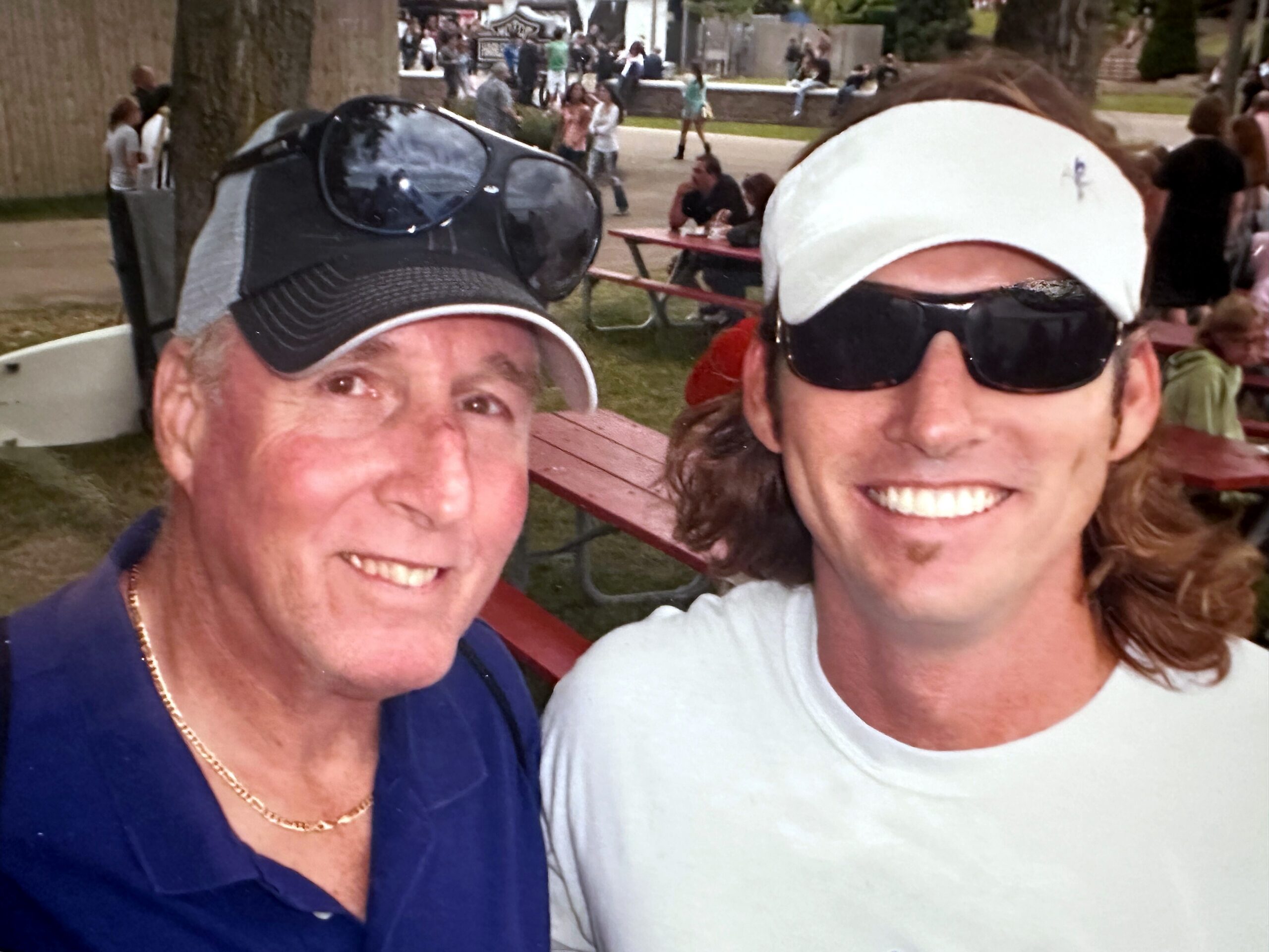 Two smiling people taking a close-up selfie at an outdoor park gathering with others in the background.