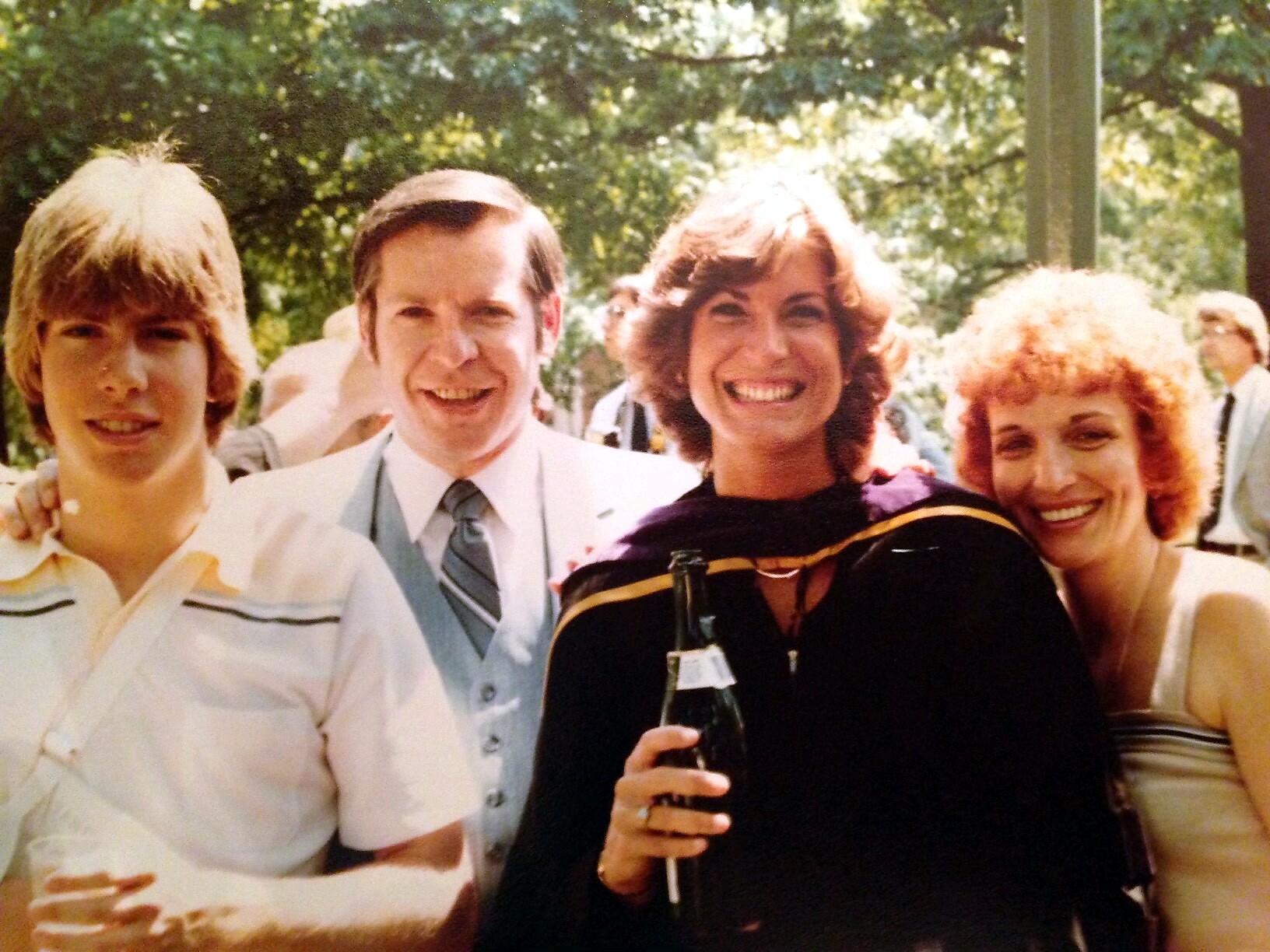 Four friends posing outdoors at a celebration; the woman in black holds a beer bottle and smiles broadly.