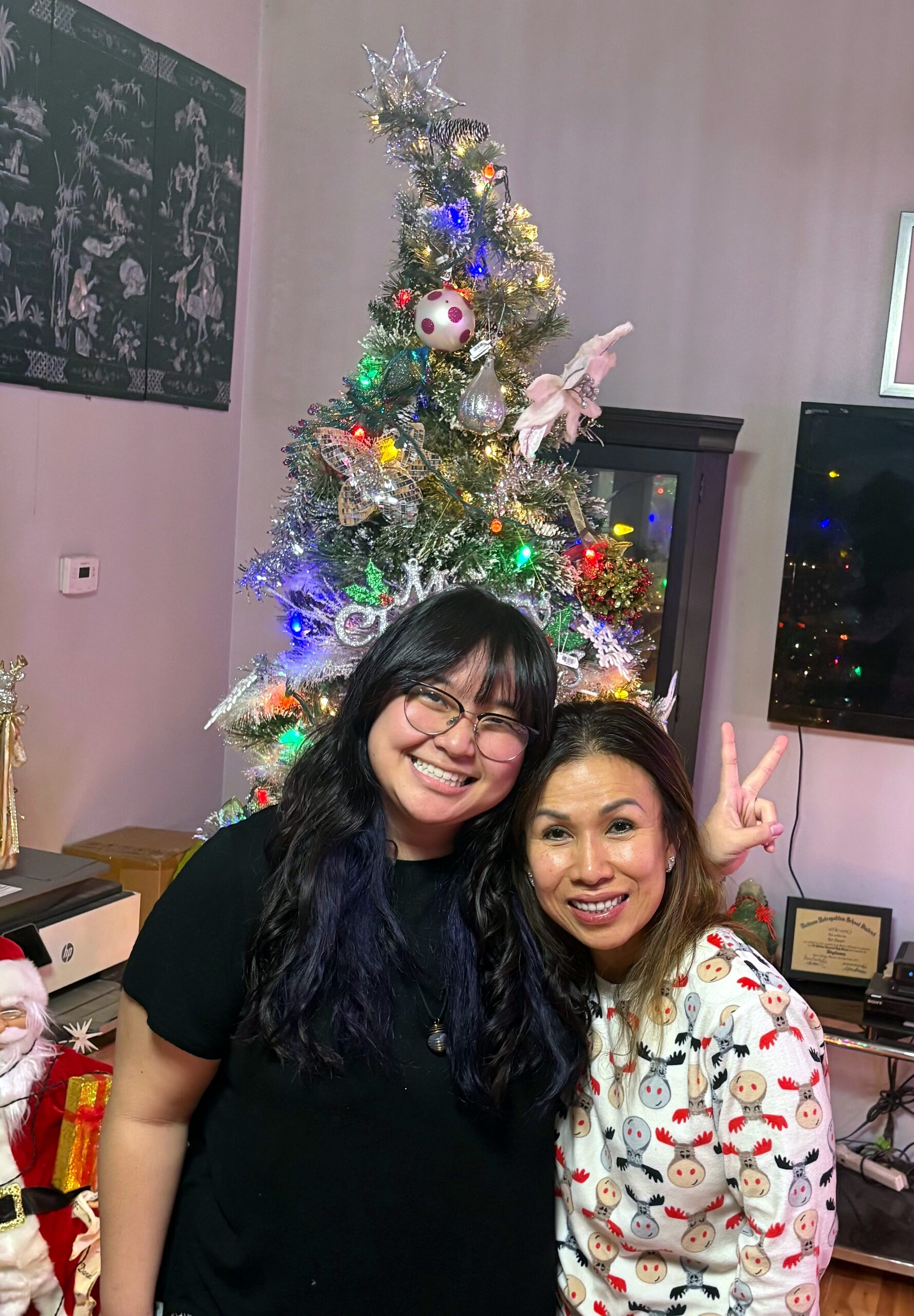 Two smiling women posing in a living room beside a decorated Christmas tree with colorful lights and ornaments.