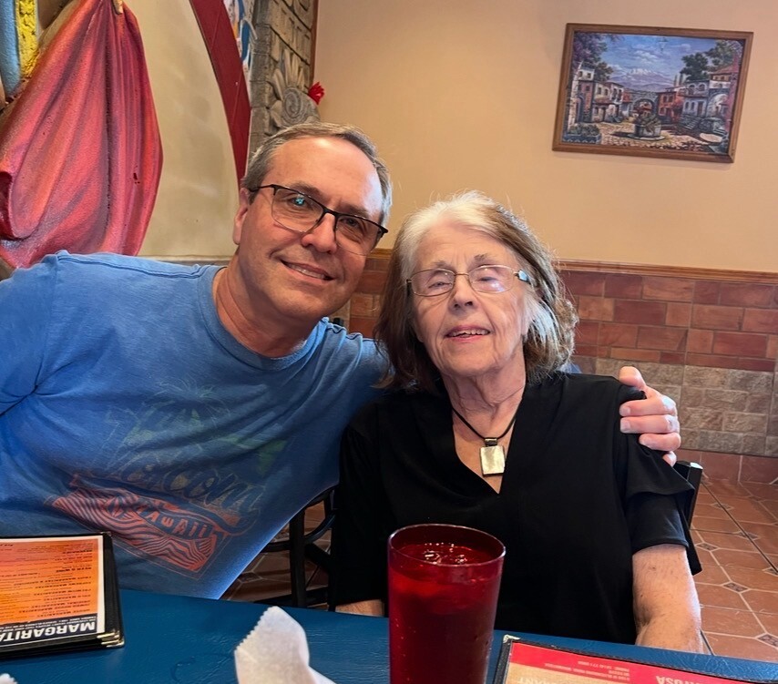 Man in a blue T-shirt with glasses sits beside a smiling elderly woman in a black top at a restaurant table with a red drink nearby.
