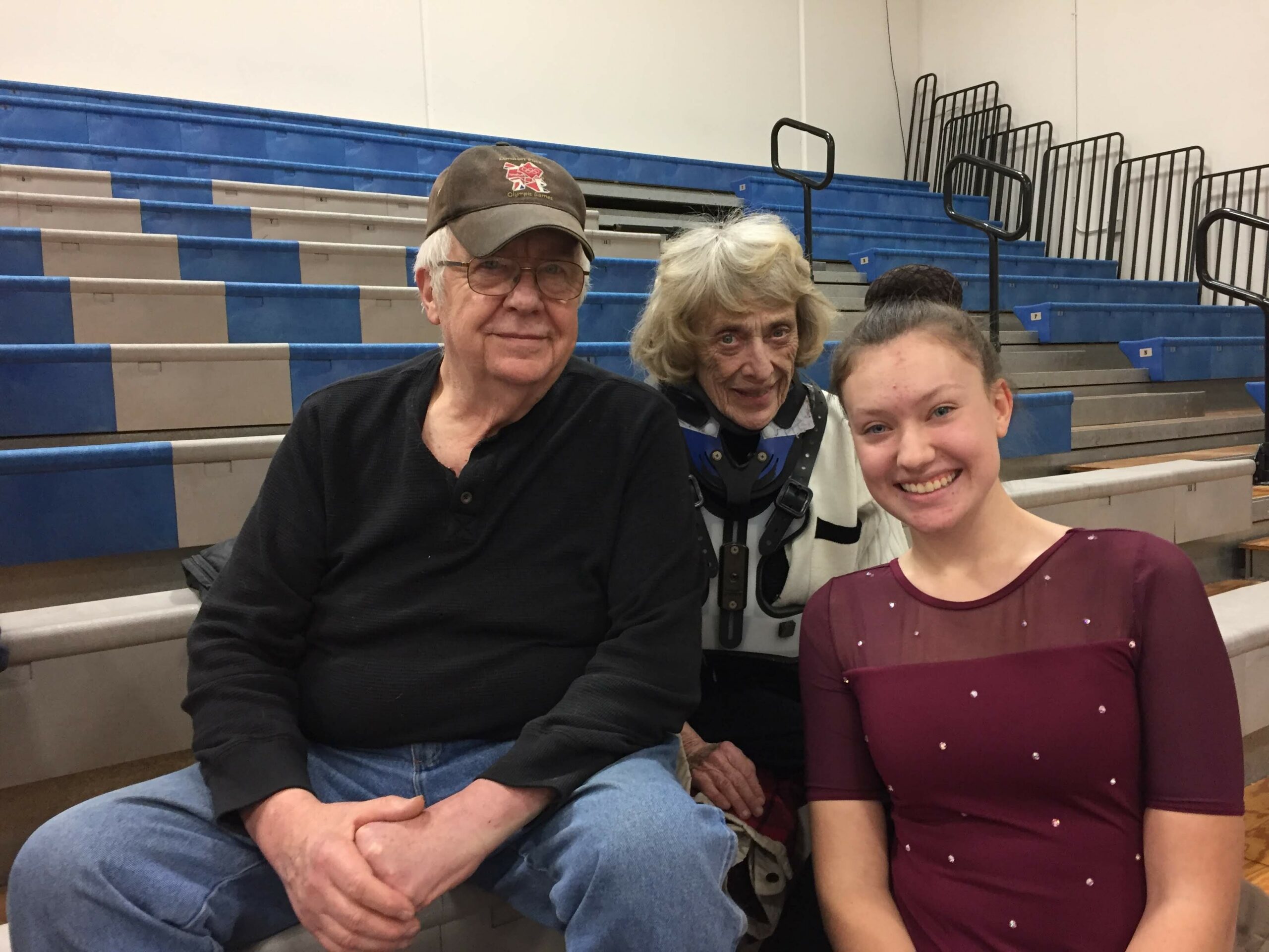 Three people pose for a photo on gym bleachers: a man with a cap on the left, an older woman in the center, and a smiling young woman in a maroon top on the right.