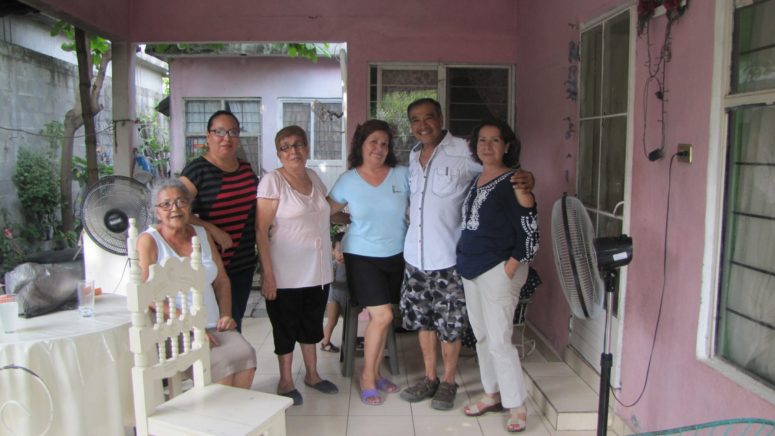 Six adults pose together on a pink patio with a white tiered cake on a table in front of them.
