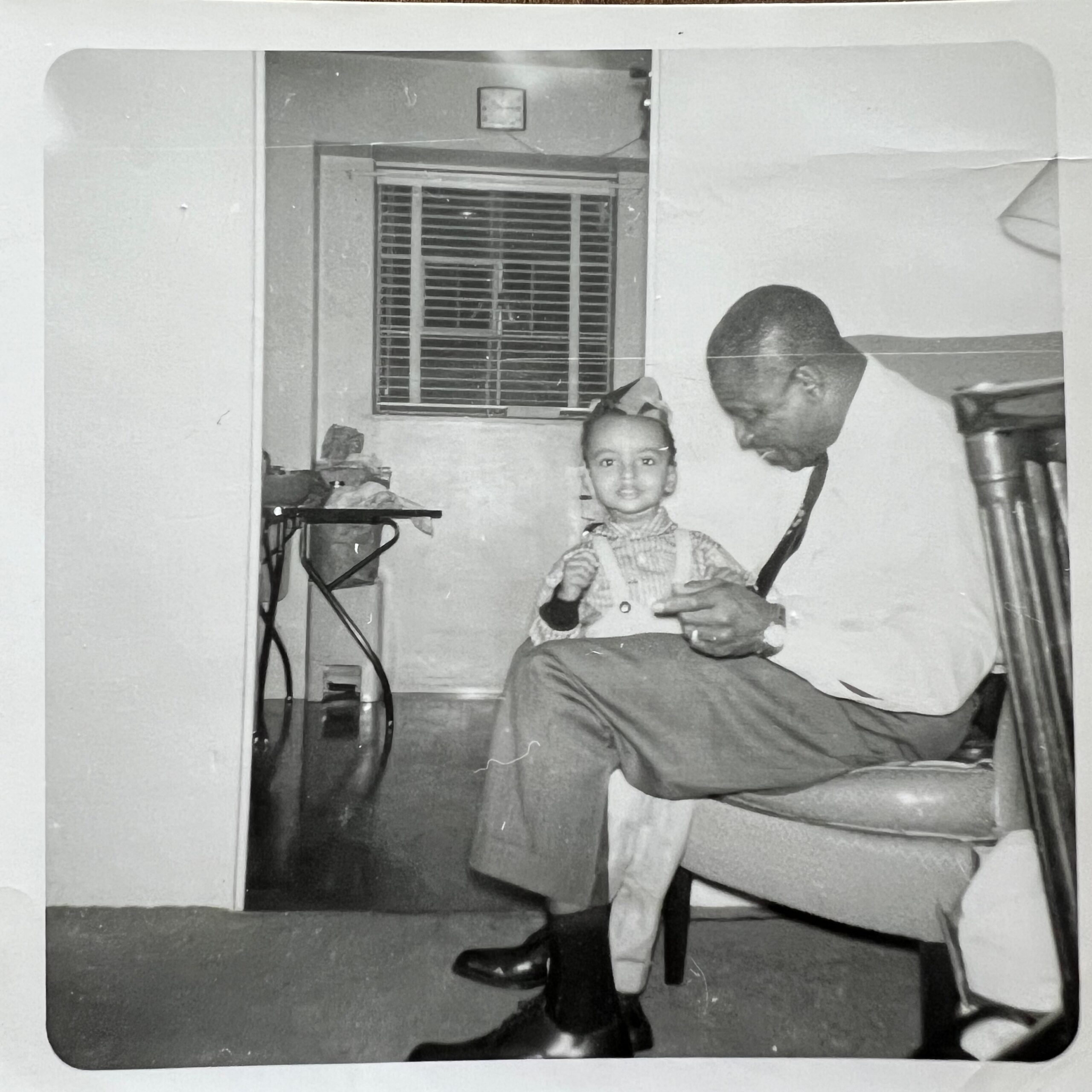 Child in plaid shirt stands in a living room beside a seated man, with a window and blinds behind them.