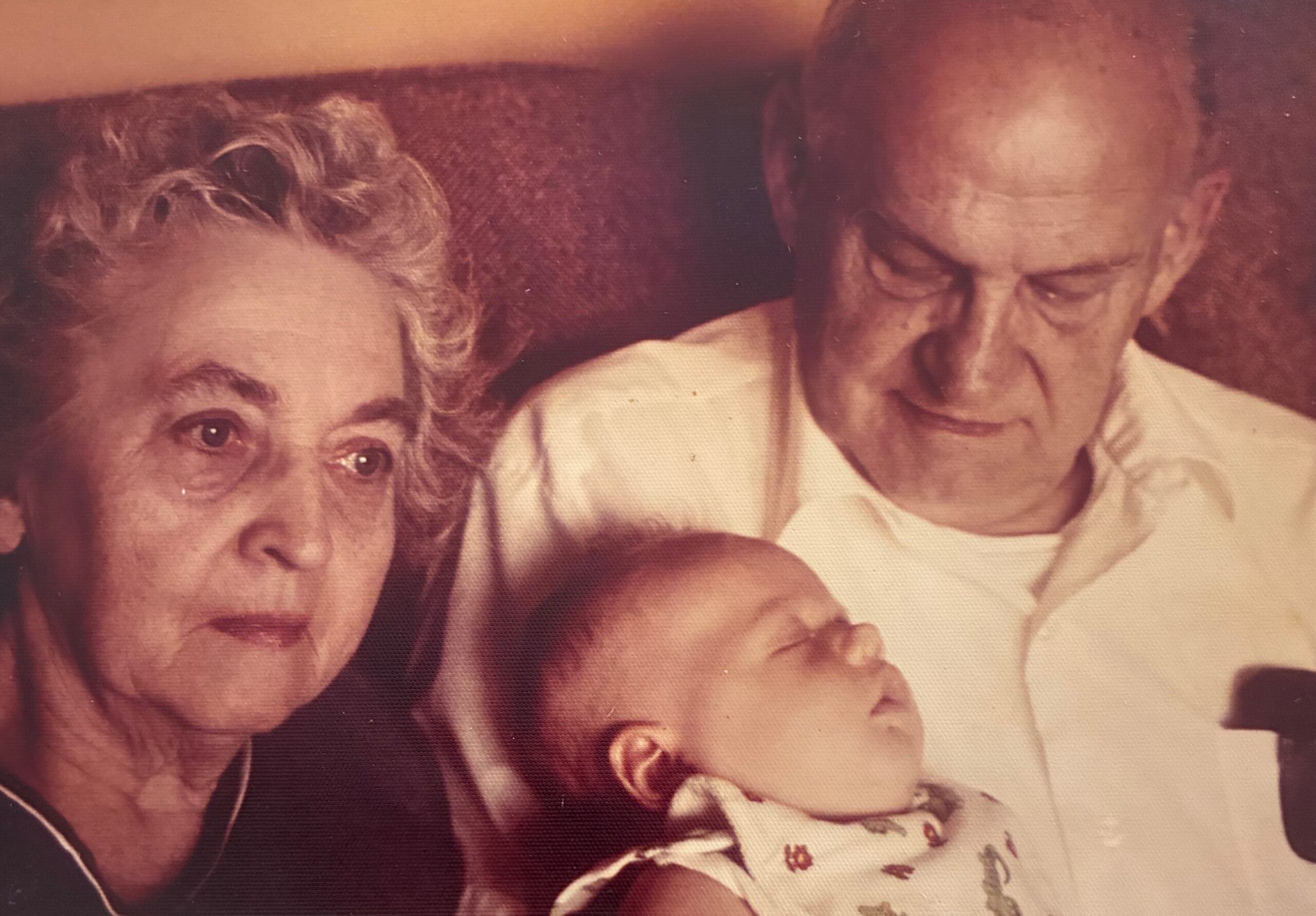 Close-up of an elderly woman with short gray hair and an elderly man leaning in beside a baby between them, in a warm, grainy photo.
