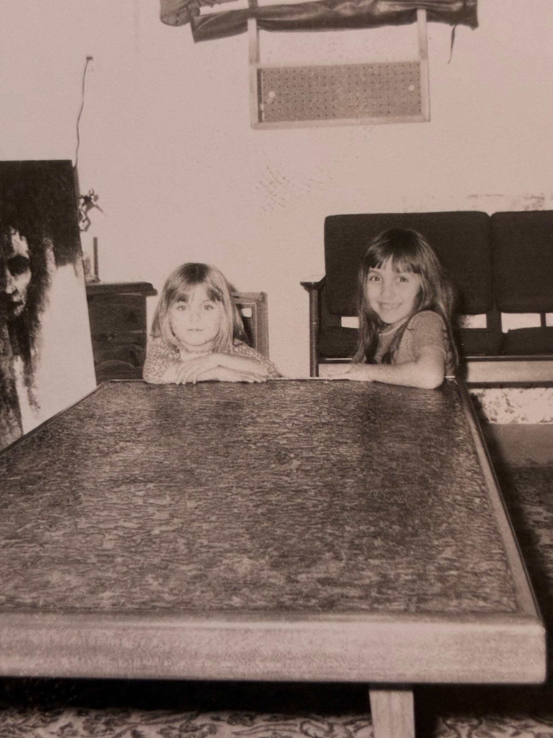 Two young girls sit at a low table or bed in a living room, smiling at the camera.
