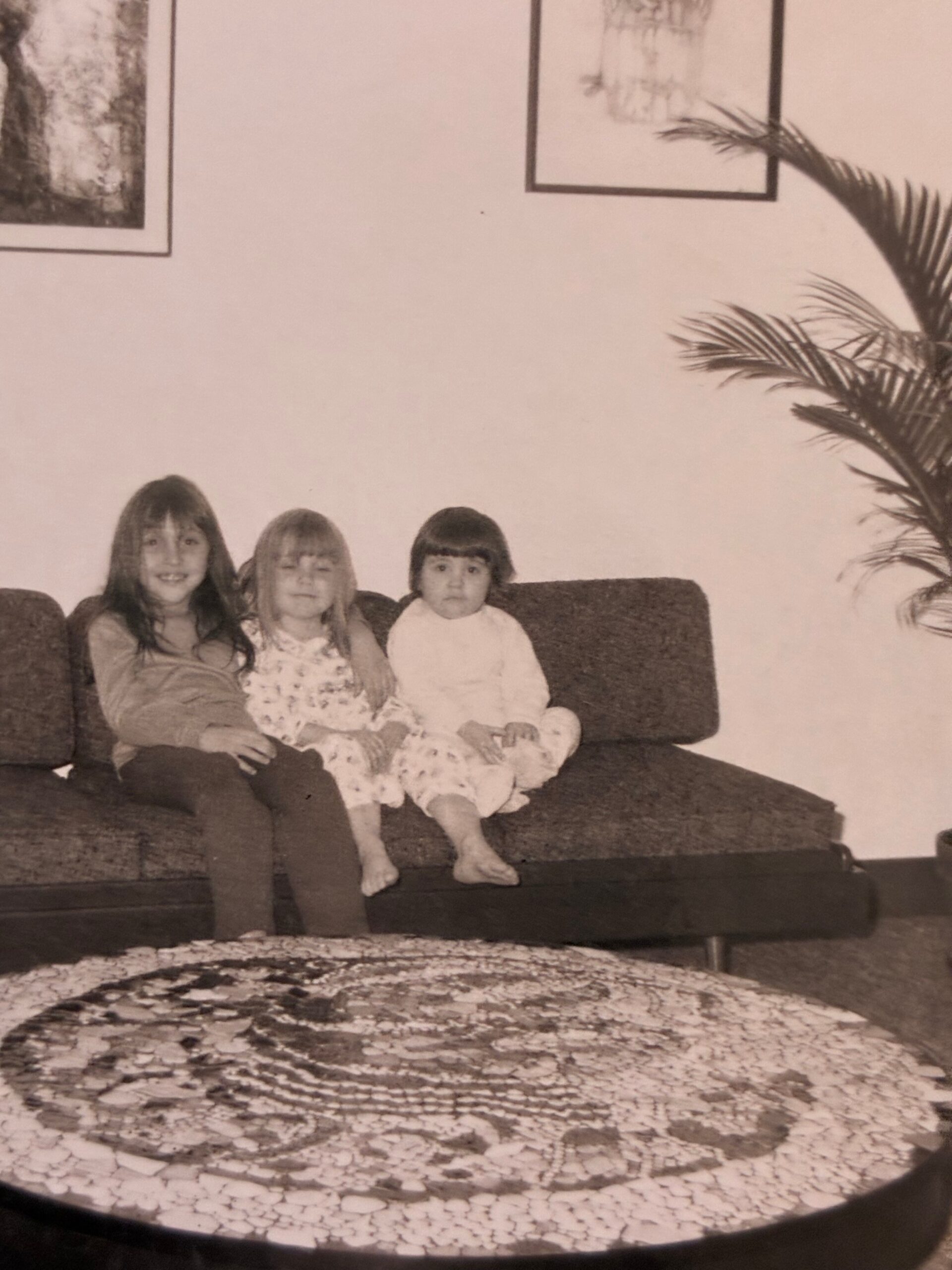 Three children sit together on a dark couch in a living room, with a palm plant to the right and framed pictures on the wall behind.