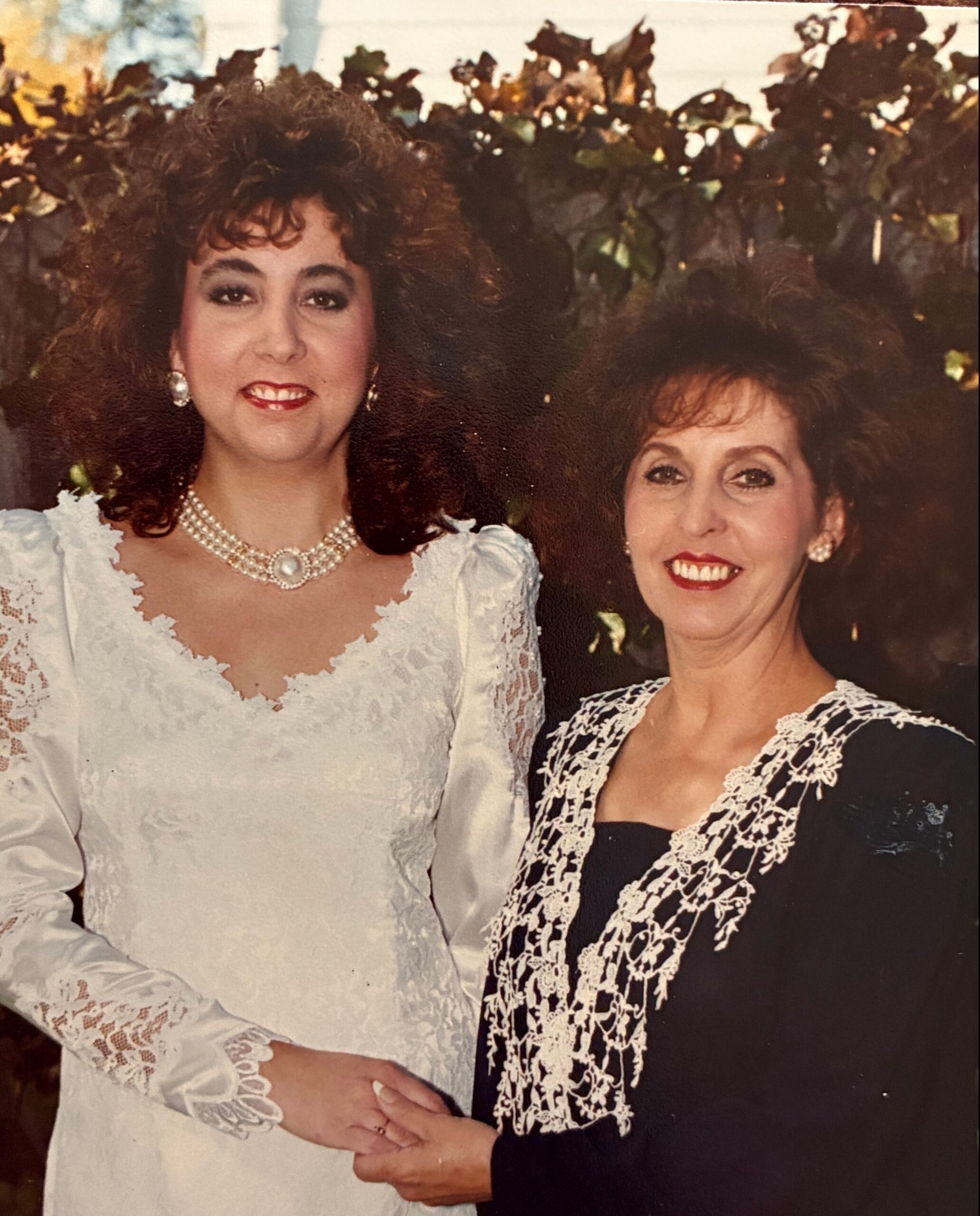 Two women in wedding attire pose together, one in a white lace dress with pearls, the other in a black dress with lace trim, outdoors behind them.