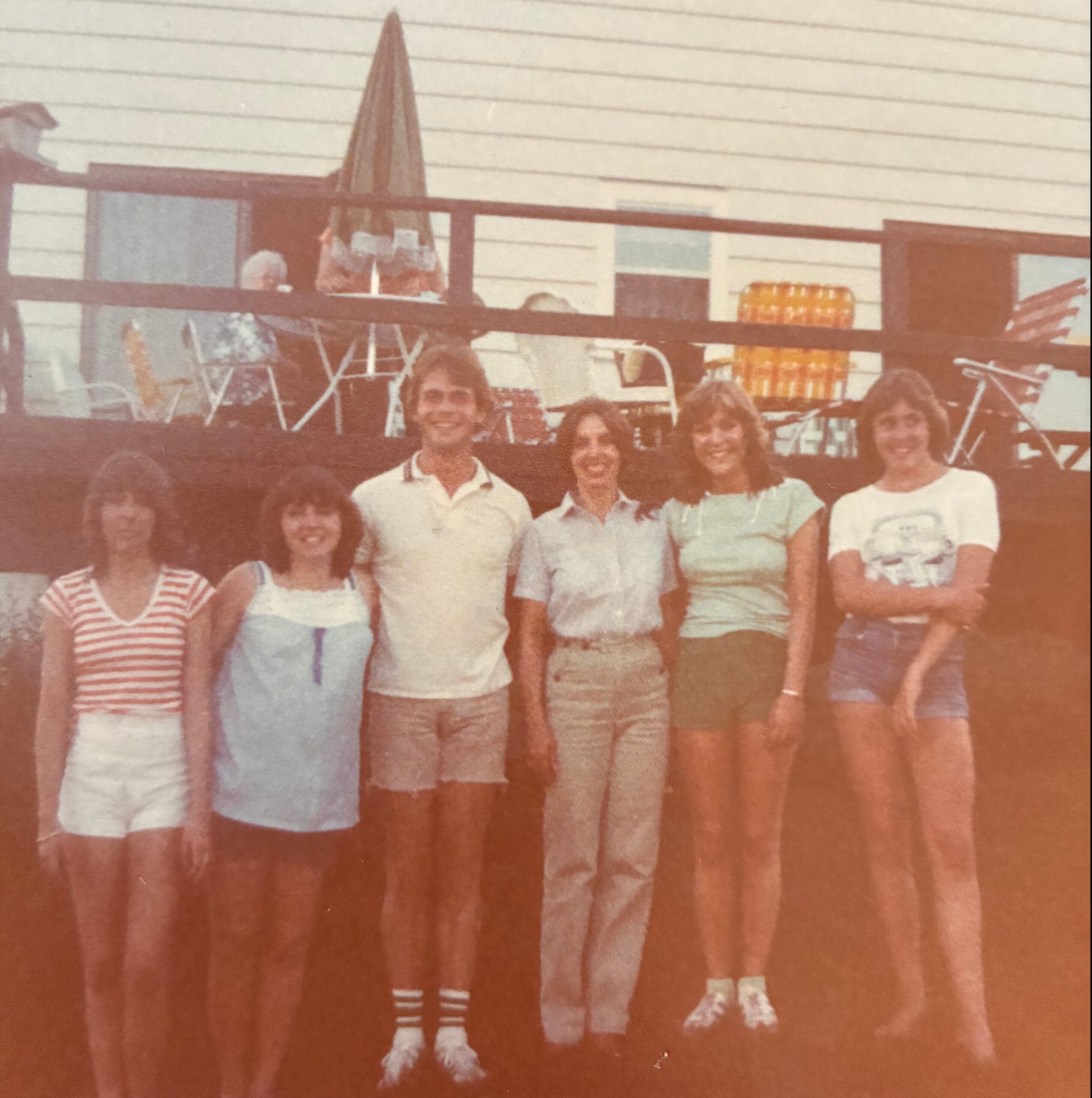 Group of young adults posing together on a wooden deck with a railing, outdoors on a sunny day.