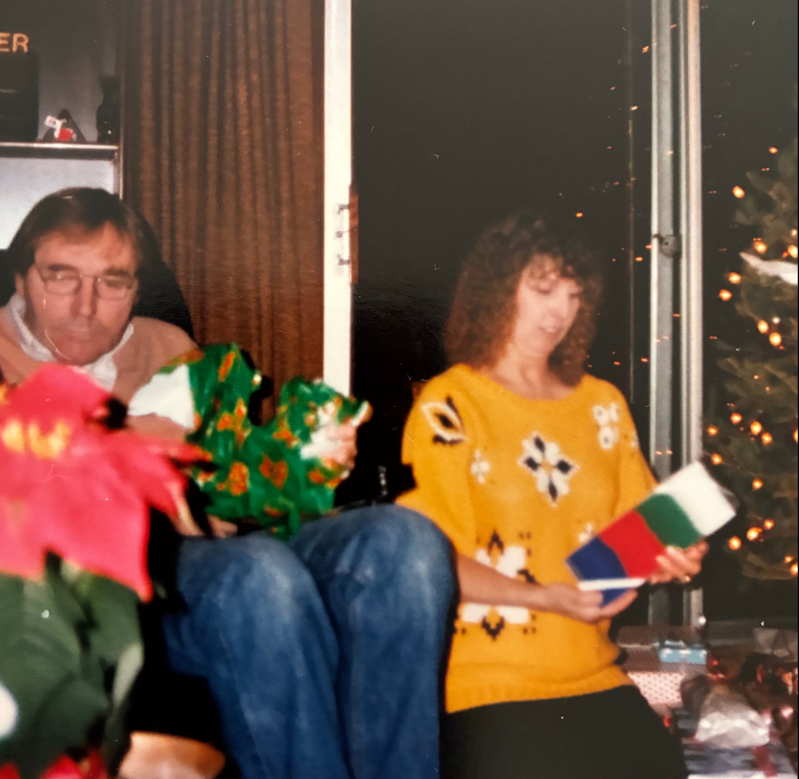 People at a holiday party wearing colorful sweaters, standing near a window with Christmas lights in the background.